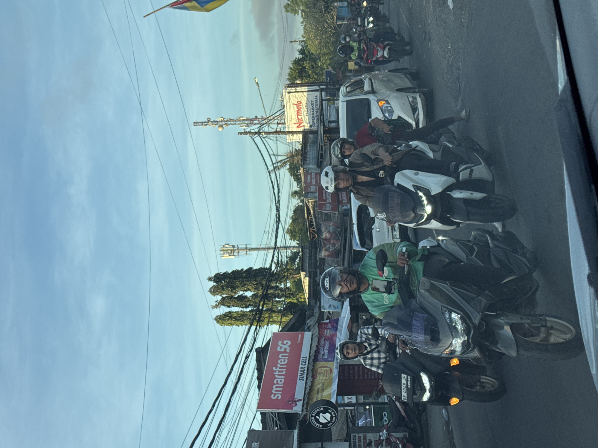 The image captures a street scene in Kuta Selatan, Indonesia, characterized by dense traffic primarily composed of motorcycles. Several individuals are seen riding motorbikes, most wearing helmets. Some appear to be Grab drivers, based on the visible Grab logo on their attire. Street-side advertisements are prominent, including a large "smartfren 5G" sign and a "Nirmala Pecatu" supermarket sign. Overhead power lines are numerous, and telecommunication towers are visible against a blue sky. The scene reflects the typical urban environment of Kuta Selatan with its bustling activity and mix of commercial and infrastructure elements.