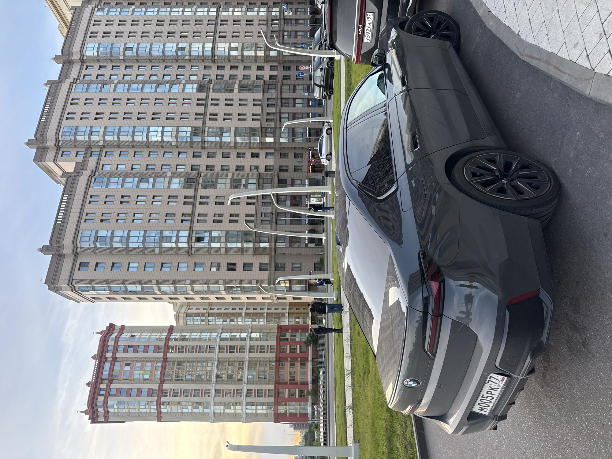 A gray BMW automobile is parked on a street in Moscow, Russia. The building facades in the background are visible. There are a few people nearby. The vehicle has a license plate from Russia. The environment is urban. The interaction between people appears casual.