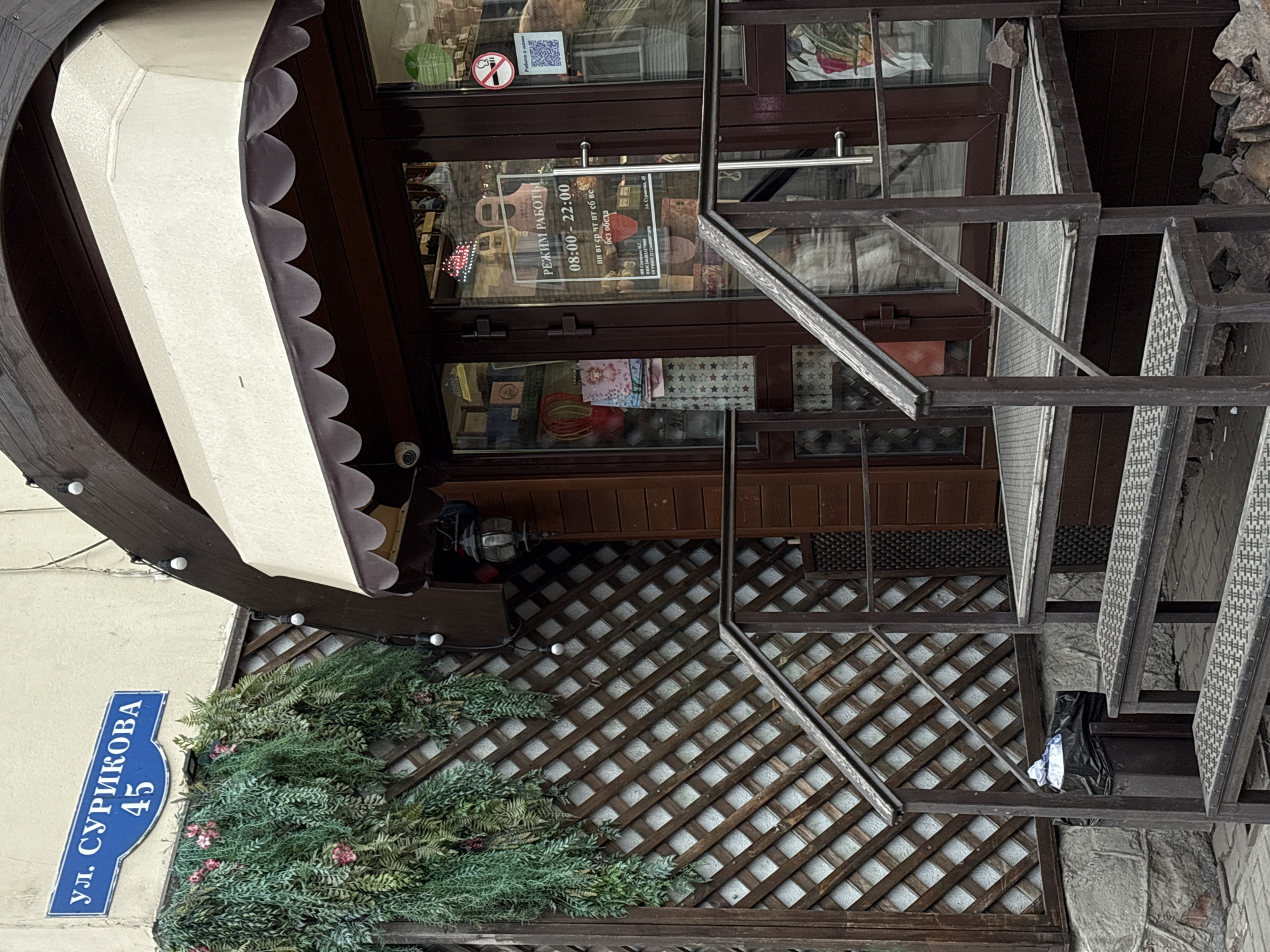 Here is a detailed description of the image:
**Scene:** The image shows the entrance of a building at "ул. СУРИКОВА, 45". The building likely has a commercial purpose, given the "РЕЖИМ РАБОТЫ" (operating hours) sign in the window, indicating it's open from 08:00 to 22:00 daily without lunch breaks.
**Objects and Details:**
* **Building Entrance:** The entrance consists of a brown-framed glass door with a dark wood trim. Above the door is an awning with a white canopy and decorative scalloped edges.
* **Street Sign:** A blue street sign displaying "ул. СУРИКОВА, 45" is affixed to the building.
* **Vegetation:** There's decorative greenery, possibly artificial, arranged on a lattice-patterned facade.
* **Staircase:** A metal staircase leads to the entrance.
* **Signage:** There's an opening hours sign on the door, along with no smoking sign and a QR code.
**Location:** Based on the street name, the location is likely in the city of Krasnoyarsk, Russia.