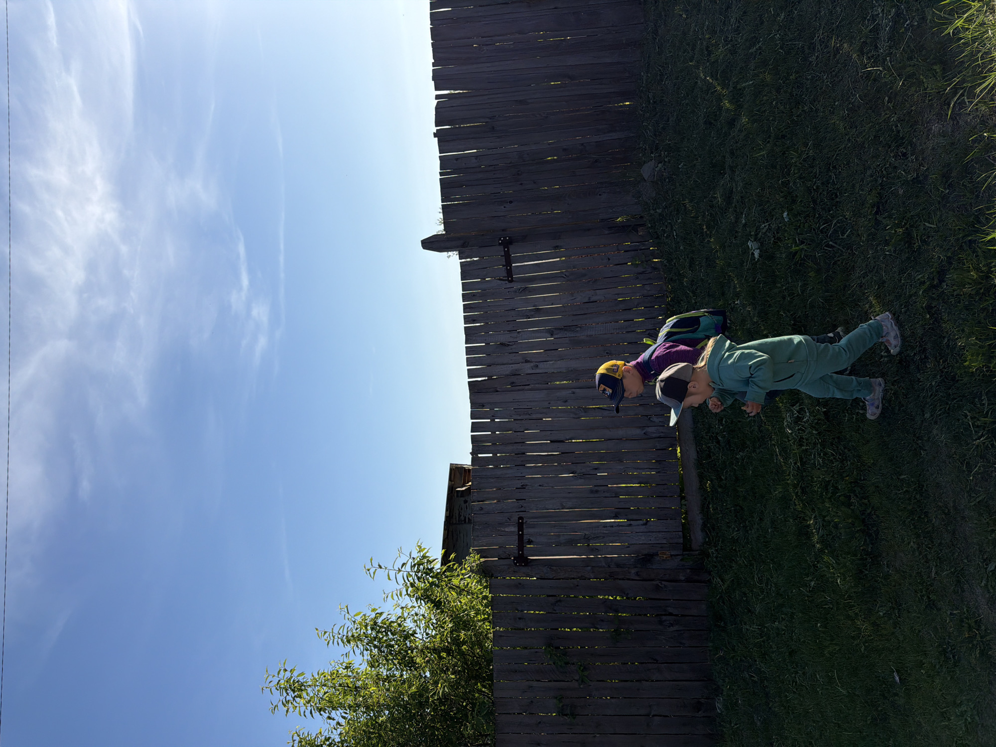Two children are near a wooden fence. One child wears a purple shirt and a baseball cap, and the other wears a green outfit and a baseball cap. There is grass and green foliage near the fence. The sky is blue with light clouds. The location is outdoors and appears to be a yard or field. The specific location in the city of Chaikovsky, Russia, cannot be determined from the image.