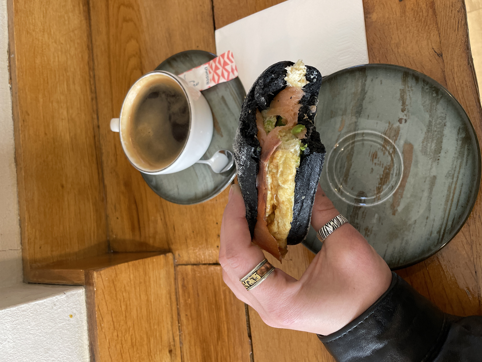 The image shows a person holding a black sandwich, presumably a charcoal bread sandwich, with visible fillings of scrambled eggs, salmon, and green vegetables (possibly sprouts or arugula). The sandwich is partially eaten. The person is wearing a black leather jacket and two rings on their fingers: one with a gold and copper design, the other silver with a textured pattern.
In the background, there is a cup of black coffee on a saucer and a packet of sugar on a table with a light-colored napkin nearby. A second empty plate is also visible. The table surface is wooden with a warm, natural finish.
It is impossible to verify location in Tbilisi. The scene suggests an indoor café or restaurant setting.