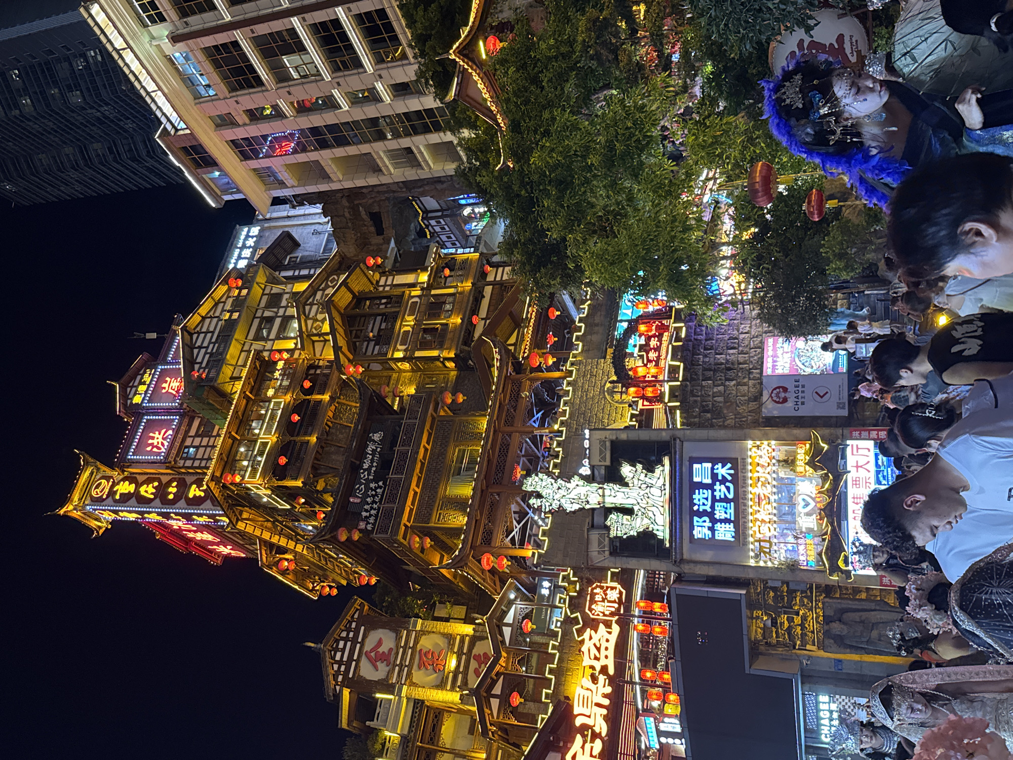 The image shows a nighttime scene in the Yuzhong District of Chongqing, China, with a focus on a brightly lit, multi-story traditional Chinese-style building adorned with red lanterns. The architecture is intricate, with layered roofs and wooden balconies. Neon signs in Chinese characters illuminate the facade, and a prominent vertical sign displays blue and white text. To the right of the traditional building, a modern skyscraper with numerous windows is visible against the dark sky. In the foreground, a crowd of people are gathered, some dressed in elaborate costumes resembling traditional Chinese attire. One person in the foreground wears a striking blue feather headdress and ornate makeup. The overall atmosphere is vibrant and festive, suggesting a lively urban environment at night.