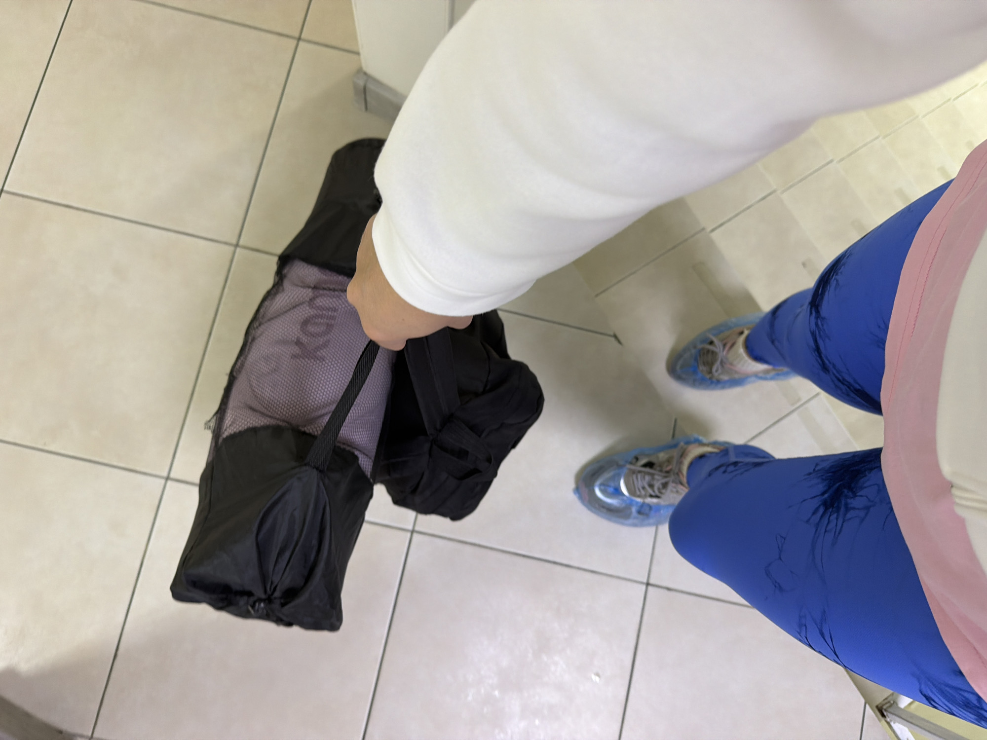 The image shows a person holding a black bag with gray mesh in a tiled area. The person is wearing blue leggings, a white sweatshirt, and gray and white sneakers covered in blue shoe covers. The tiles are light beige or cream colored. The location in the city of Sochi, Russia, cannot be determined based on this image alone.