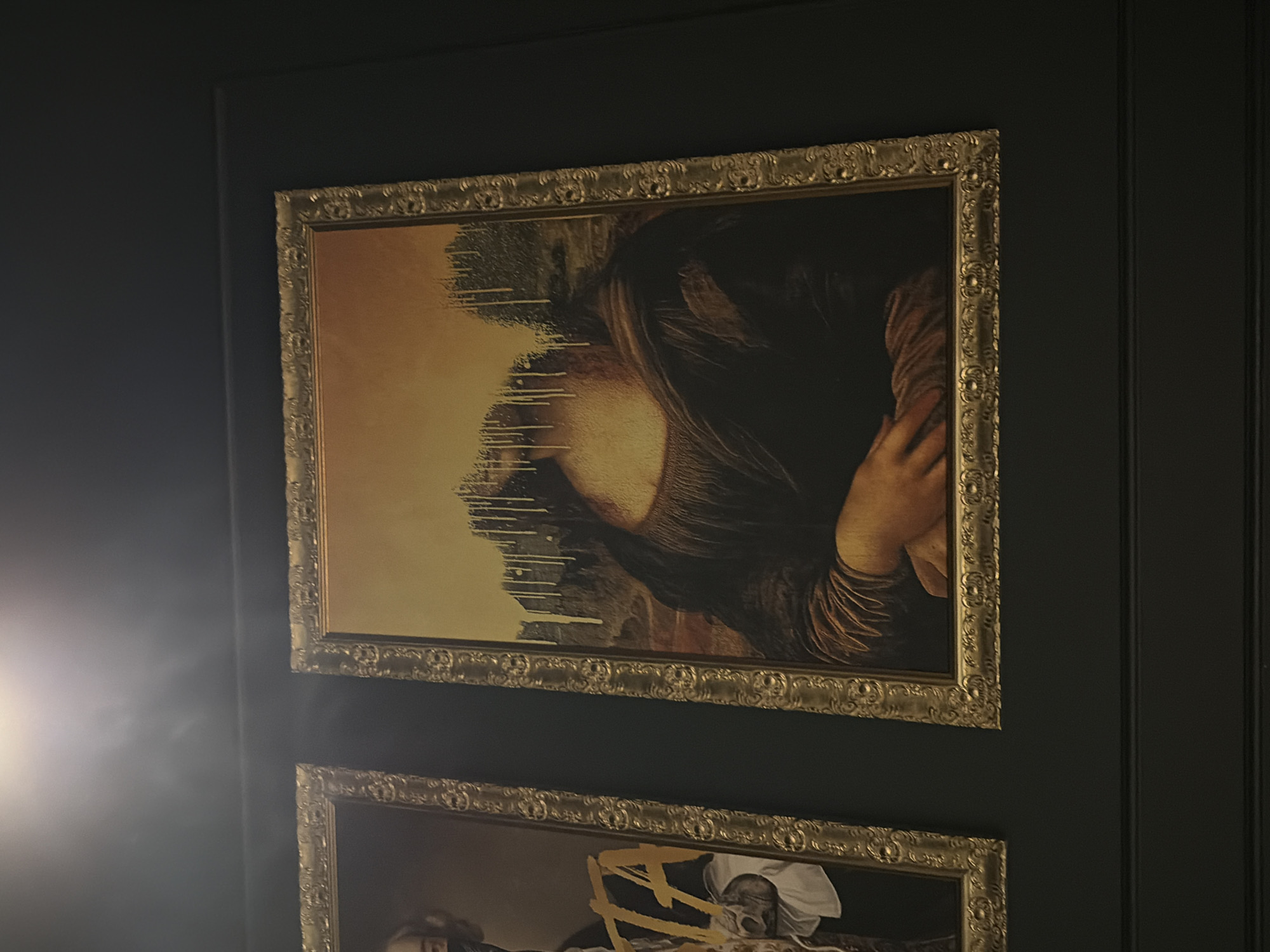 A close-up shot reveals two framed artworks hung on a dark, possibly black, paneled wall. The top artwork features a modified image of Leonardo da Vinci's Mona Lisa. The left side of the portrait has been altered to appear as though it is melting or dissolving, with streaks running down the canvas. The artwork below this one is partially visible, showing at least two figures and a white cloth or garment. Both artworks are framed in ornate, gold-colored frames. The scene is dimly lit, with a light source appearing from the left side of the frame, casting a subtle glow. No specific location in Chaikovsky, Russia is identifiable from the visual information provided.