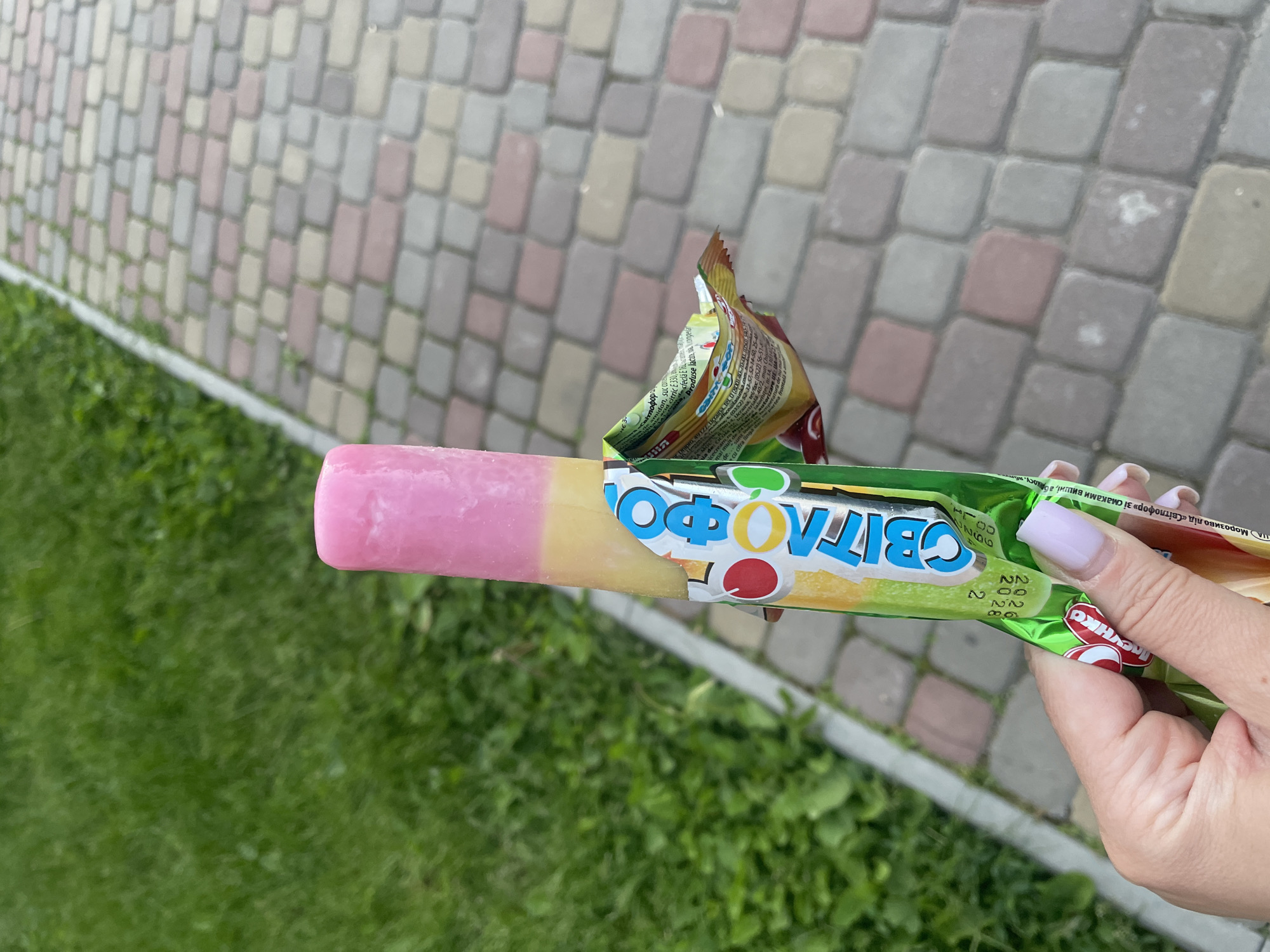 A close-up, high-angle shot shows a person's hand holding a multi-colored ice cream popsicle outdoors. The ice cream itself has distinct pink and yellow layers, suggesting different fruit flavors. Its green wrapper is partially torn open, revealing the product, and features the brand name "Світлофор" (Svitlofor, meaning "Traffic Light") prominently in Cyrillic script with colorful circular elements.
The hand, likely female given the manicured nails with light lavender polish, grasps the wrapper, which also displays smaller text. Readable snippets of this text include "Морозиво лід «Світлофор» зі смаками вишні, ананасу" (Ice cream "Svitlofor" with cherry and pineapple flavors), "Ucraina" (Ukraine), and contact numbers, indicating the product's origin.
The background consists of a textured pavement made of interlocking rectangular blocks in varying shades of grey, beige, and light reddish-brown, stretching away into the distance and slightly out of focus. To the left and below the ice cream, vibrant green grass is visible, suggesting a natural border alongside the paved area. The bright, even lighting indicates it is daytime, likely sunny, providing a clear view of the scene. The overall setting implies a casual outdoor moment, possibly on a sidewalk or path in Rozsoshentsi, Ukraine.