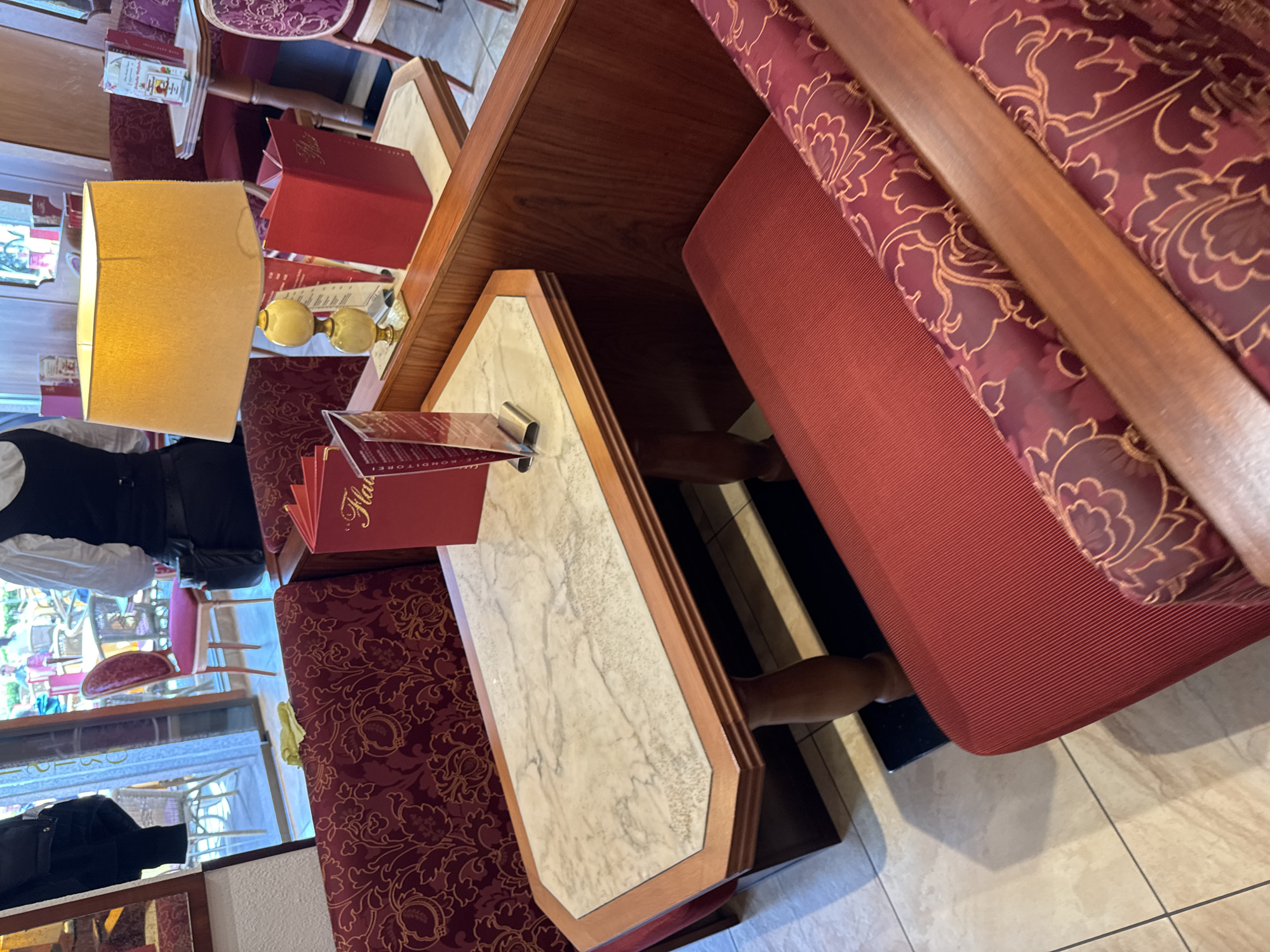 The image shows the interior of a café, likely in Cochem, Germany. A table with a marble top and a wooden frame is partially visible in the foreground. On the table is a menu in a red holder. The café seating includes a red padded booth. A lamp with a yellow shade is present. A waiter wearing a white shirt and black vest is partially visible. In the background, there are other tables, chairs, and patrons. The floor is tiled in light colored. The overall setting suggests a traditional European café environment.