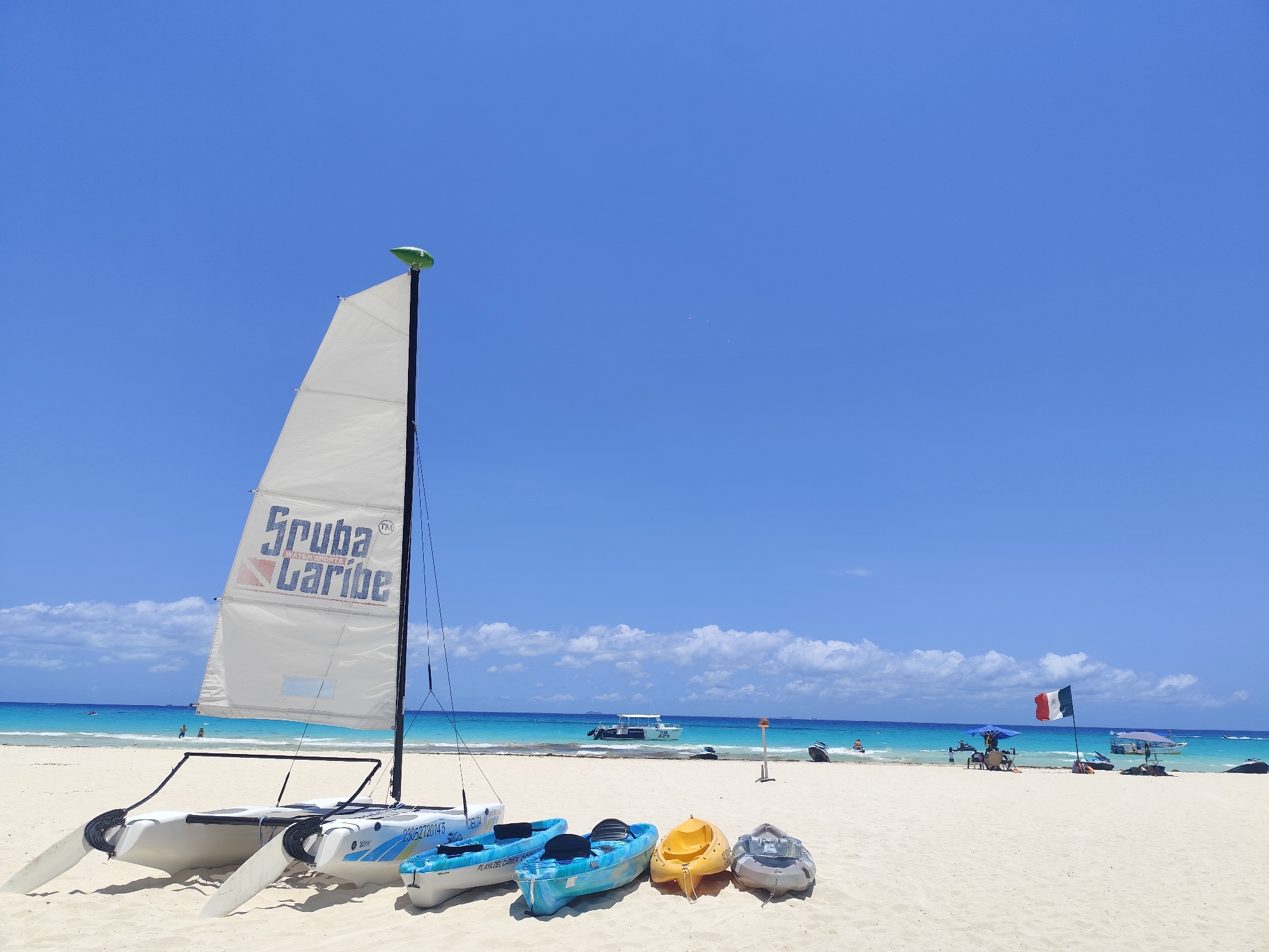The image captures a sunny beach scene in Playa del Carmen, Mexico. In the foreground, a catamaran sailboat with a white sail emblazoned with "Scuba Caribe" sits on the white sand. Next to the catamaran, four kayaks of varying colors (blue, yellow, and gray) are arranged.
In the background, the turquoise water of the Caribbean Sea meets the beach. Several boats are visible on the water. A Mexican flag waves in the gentle breeze. Some people can be seen swimming and relaxing near the shore. An umbrella provides shade for beachgoers in the distance. The sky is a clear, vibrant blue with a few scattered clouds.