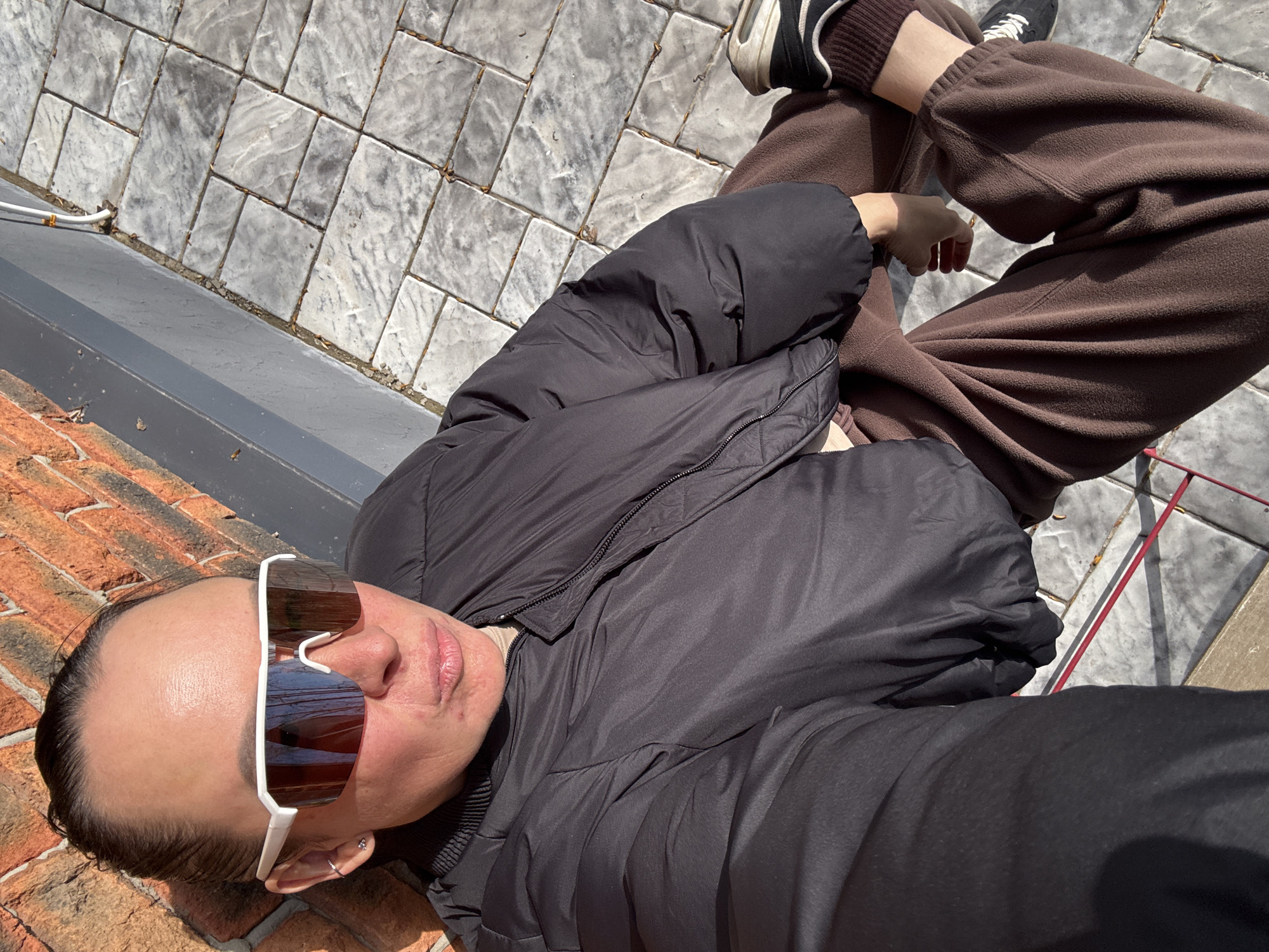 A person wearing sunglasses and a black puffer jacket lies on a brick and concrete surface. The person also wears brown sweatpants and black and white sneakers. The setting is outdoors, possibly a patio or sidewalk in Chaikovsky, Russia, based on the information provided. The person seems to be looking upwards, possibly taking a selfie or resting.