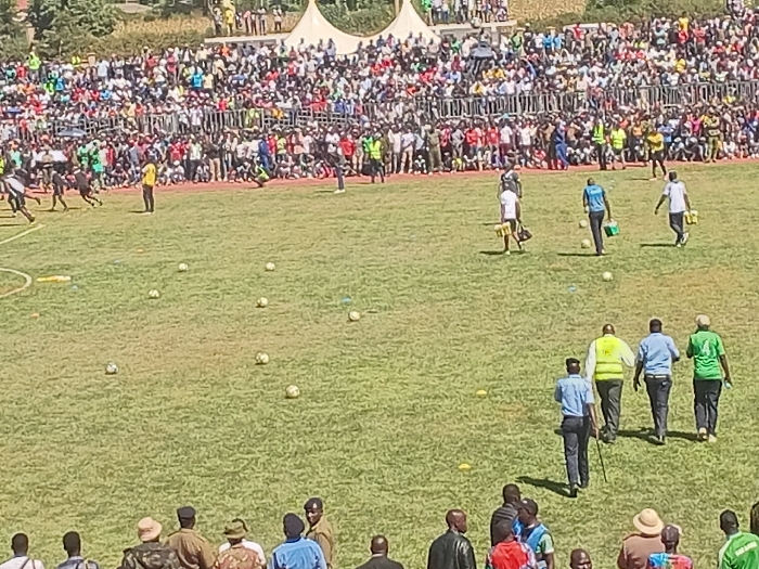 The image depicts an outdoor football (soccer) event taking place in a stadium under bright daylight. A wide, green grassy pitch dominates the foreground and midground, scattered with numerous footballs and small yellow marker cones.
In the background, tiered stands are densely packed with a large crowd of spectators, indicating a significant public gathering. Behind the main crowd, white event tents are visible, with distant hills and trees forming the horizon.
On the field, several individuals are active. To the left, a group of people, possibly players in dark sportswear, are in motion, with one person in a yellow shirt standing nearby. In the center, four individuals in light-colored shirts (white and blue) are seen walking, some appearing to carry equipment or water bottles. Further in the mid-ground, a group of four men walks across the field: one wears a high-visibility yellow vest, two are in light blue shirts, and one is in a green shirt. One of the men in a blue shirt holds a stick or cane. In the lower foreground, several other men, some possibly in uniform, are seen from behind, observing the activities on the pitch.
The presence of many balls, active participants, and a large audience suggests a practice session, a warm-up, or a community football match or tournament. The bright sun indicates a clear, sunny day, conducive to outdoor sports. The overall atmosphere appears lively and engaging, consistent with a community event in Siaya, Kenya. No discernible text is visible in the image.