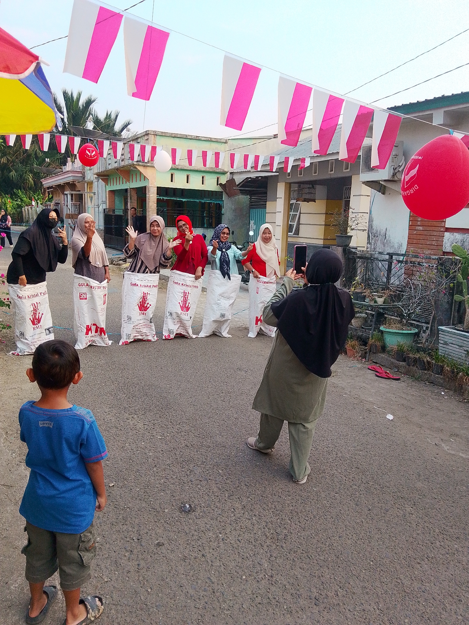 In the city of Barabai, Indonesia, an outdoor street scene captures a festive atmosphere with a sack race in progress. Five women, all wearing headscarves, stand with their lower bodies inside large white sacks, suggesting they are participating in the race. They are positioned on an asphalt road, with houses and a fence line visible in the background. One woman in the foreground, facing away from the camera and wearing a black headscarf, is actively recording the event on her mobile phone. A young boy, seen from the back in a blue t-shirt and olive-colored shorts, stands on the road, appearing to watch the proceedings. Overhead, white and pink pennant banners are strung across the street, along with a large red balloon and a smaller white balloon, adding to the celebratory ambiance. The setting appears to be a residential area, with plants in pots along the fence and a partially visible umbrella in the upper left corner.