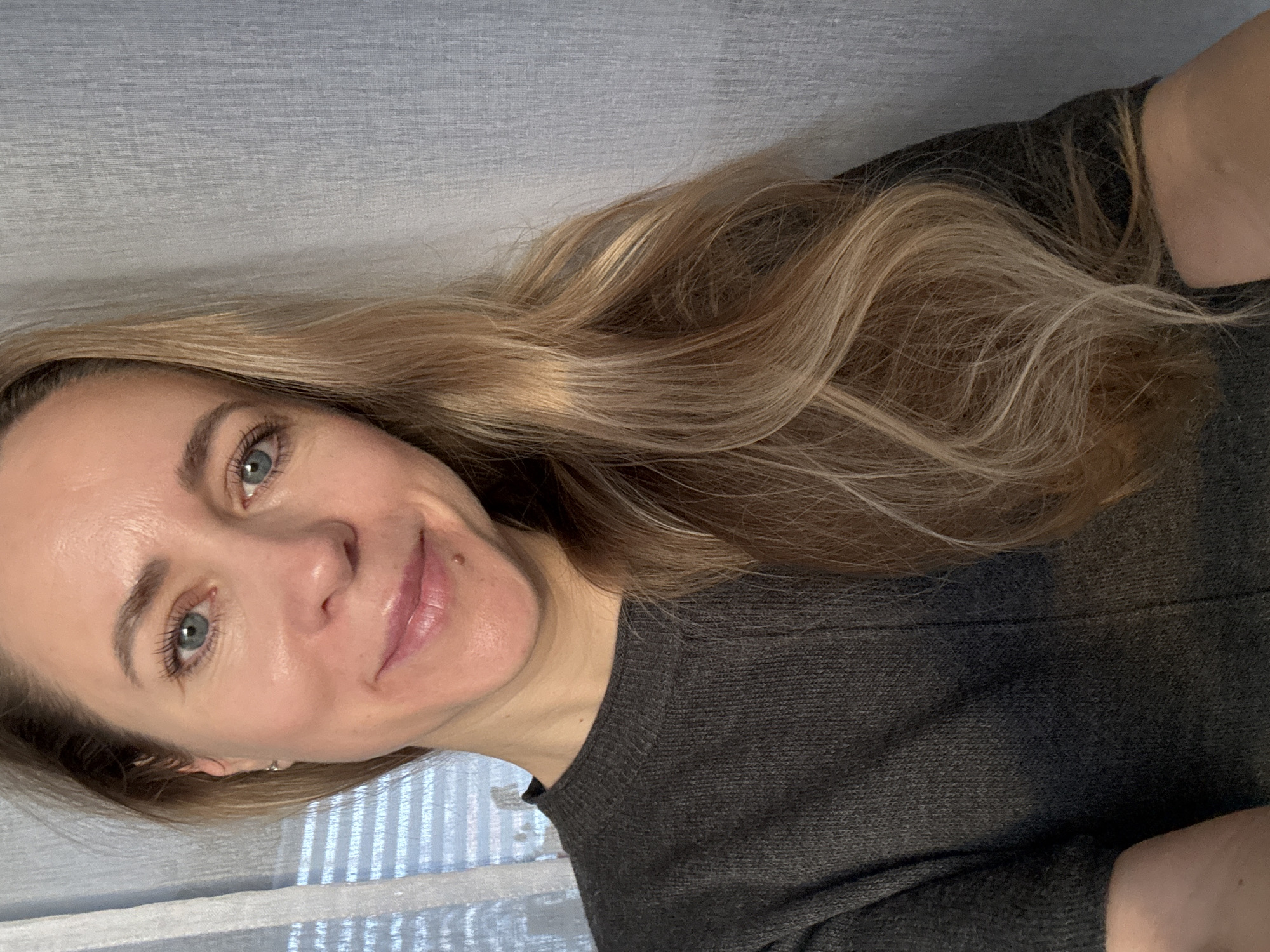 A close-up selfie depicts an adult woman with long, wavy blonde hair with lighter highlights, cascading over her right shoulder. She has striking light blue eyes, a light complexion, and a small, dark mole or beauty mark positioned above her upper lip on the right side of her face. A small, subtle earring is visible in her left ear. She wears a dark grey knitted sweater with a crew neck.
The woman is looking directly at the camera with a soft, gentle smile, conveying a calm and friendly demeanor. The scene is indoors, with a light blue-grey textured surface, possibly fabric or wallpaper, forming the background behind her head and upper body. To her lower left, horizontal lines, indicative of window blinds or slats, are visible, suggesting the presence of a window and providing ambient light. The lighting appears soft and natural, consistent with an indoor setting, likely during daytime. There is no visible text in the image. The overall activity is a self-portrait.