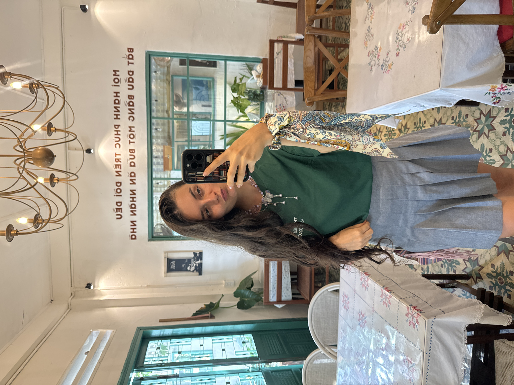 The image shows a young woman taking a selfie in a cafe. She is wearing a green t-shirt, a grey pleated skirt, and a colorful scarf tied to her wrist. She has long, dark, wavy hair and is wearing a necklace with a butterfly pendant. Her phone, in a case designed to look like a stack of books, is visible in her hand.
The cafe appears to be in Phu Thuy Ward, Vietnam. The walls are white, and there is a large window with green panes in the background. A chandelier hangs from the ceiling. Several tables with patterned tablecloths are visible, as are wooden chairs. There are plants in the background and text on the wall above the window, which appears to be Vietnamese. The flooring is tiled with a geometric pattern. The overall atmosphere is casual and inviting.
