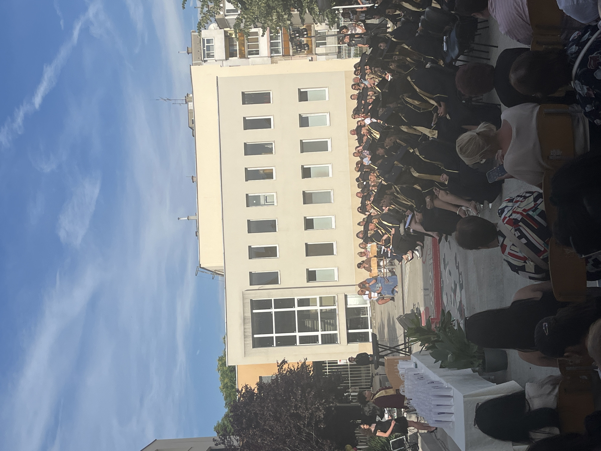 The image captures an outdoor graduation ceremony in Kazanlak, Bulgaria. The scene is dominated by a light-colored building with numerous windows, suggesting an educational institution. A large crowd of people, many dressed in academic regalia with black gowns and gold sashes, are seated and standing, indicating the event is underway or has just concluded. Some individuals appear to be family members or guests, observing the proceedings. A table covered with a white cloth is laden with what appear to be diplomas or certificates, further confirming the graduation context. In the background, trees and a clear blue sky with wispy clouds suggest a pleasant, likely daytime event. The atmosphere seems formal and celebratory, characteristic of a graduation.