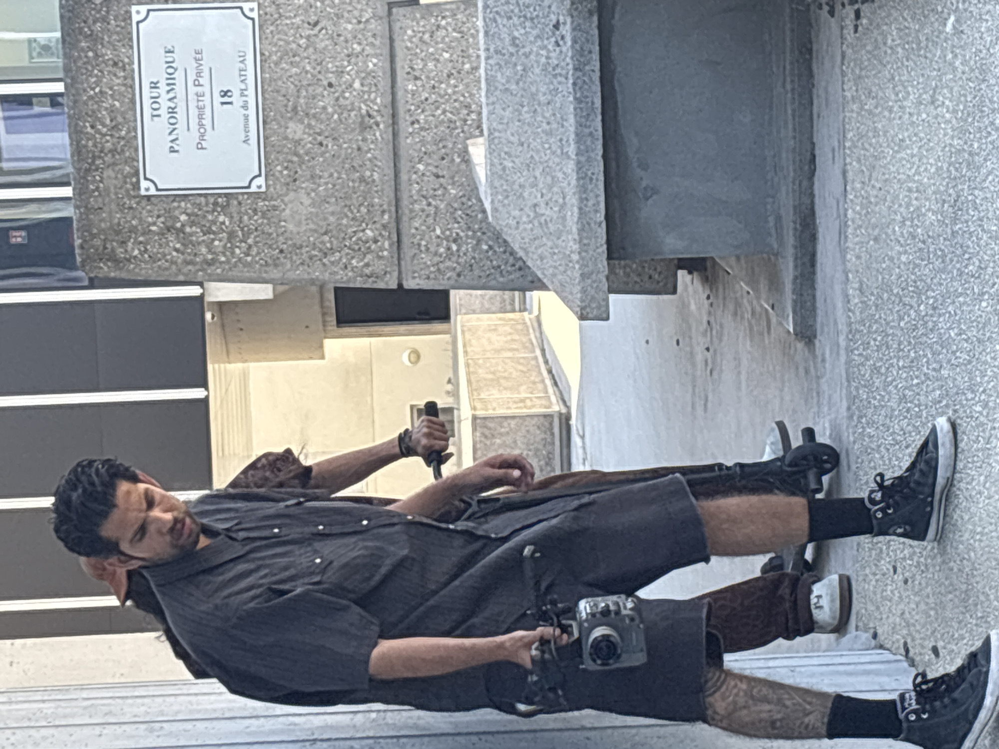 A person is lying down on a concrete ledge, possibly resting. They are wearing dark shorts, a dark button-up shirt, black socks, and black Converse sneakers. They are holding a vintage camera in their right hand, with a rig attached to it. Their left arm is extended, and they are holding a black object, possibly a flashlight or a tool. Another person is partially visible behind them, wearing a brown garment. The setting appears to be outdoors, near a building with a textured concrete facade. A sign on the building reads "TOUR PANORAMIQUE," "PROPRIÉTÉ PRIVÉE," "18," and "Avenue du PLATEAU." The scene suggests a casual, urban environment. The lighting is bright, indicating it is daytime.