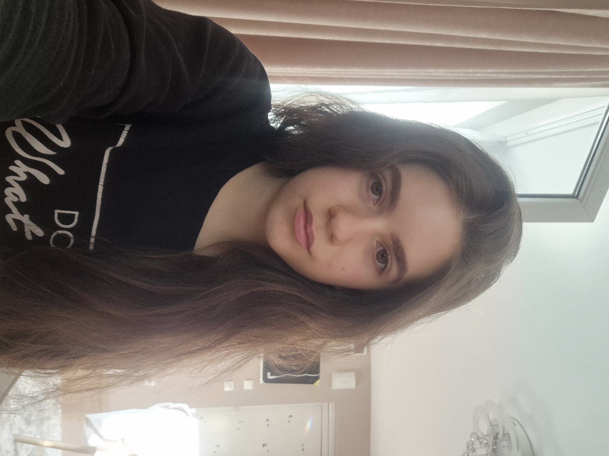 A young woman with long, dark brown hair is posing for a photo indoors. She is wearing a black long-sleeved shirt with white lettering on the front. She has fair skin, brown eyes, and full lips. The background consists of a window with beige blinds, a white wall, and what appears to be a door. The location appears to be indoors, possibly a residence. The scene is not identifiable to the city of Pavlikeni, Bulgaria.
