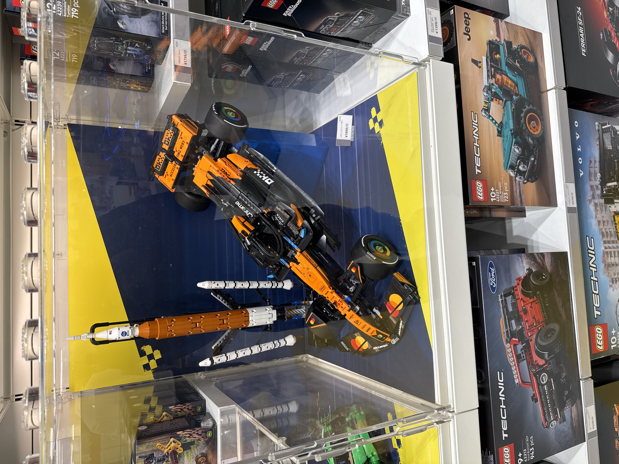 The image displays a retail store shelf featuring various LEGO Technic model car kits. The prominent display in the center showcases a large, clear acrylic case containing a LEGO model of a McLaren Formula 1 race car, predominantly orange and black with sponsor logos like "DELL" and "McLaren." Next to it, on a blue and yellow checkered background, is a LEGO model of a rocket, possibly representing the NASA Artemis I mission, with its booster stage and smaller rocket stages visible.
Below the central display, on the lower shelves, are several boxed LEGO Technic sets. These include a red LEGO Technic Ford Bronco SUV (set number 42213), a teal LEGO Technic Jeep Wrangler Rubicon (set number 42227), and other boxes with visible branding such as "Volvo" and "Ferrari SF-24." Some boxes have price tags and product numbers visible. The store appears well-lit, with white shelving and a clean, organized presentation of the merchandise. The overall impression is a modern toy store or a dedicated LEGO section within a larger retail space, likely located in a city like Liangjiang, China, given the context provided. No people are visible in the frame.