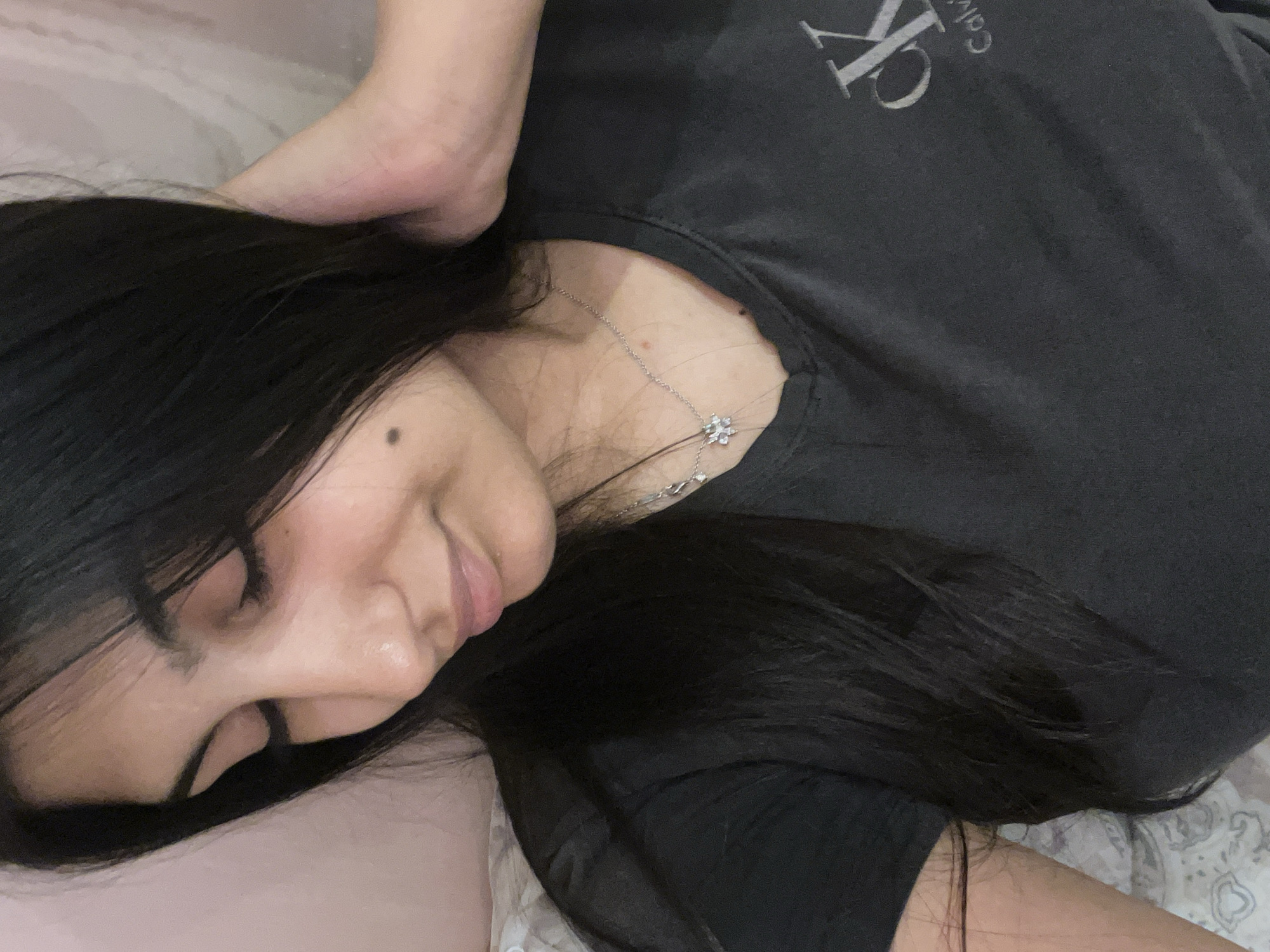A person with long, dark hair is lying down, likely indoors, as indicated by the soft lighting and domestic setting. The person is wearing a dark-colored Calvin Klein t-shirt and a delicate silver necklace with a floral pendant. The face is visible in profile, showing a closed eye and a relaxed expression. A portion of a pale-skinned limb is visible above the person's head, possibly an arm or a leg. The background appears to be bedding with a pale pink or white cover.