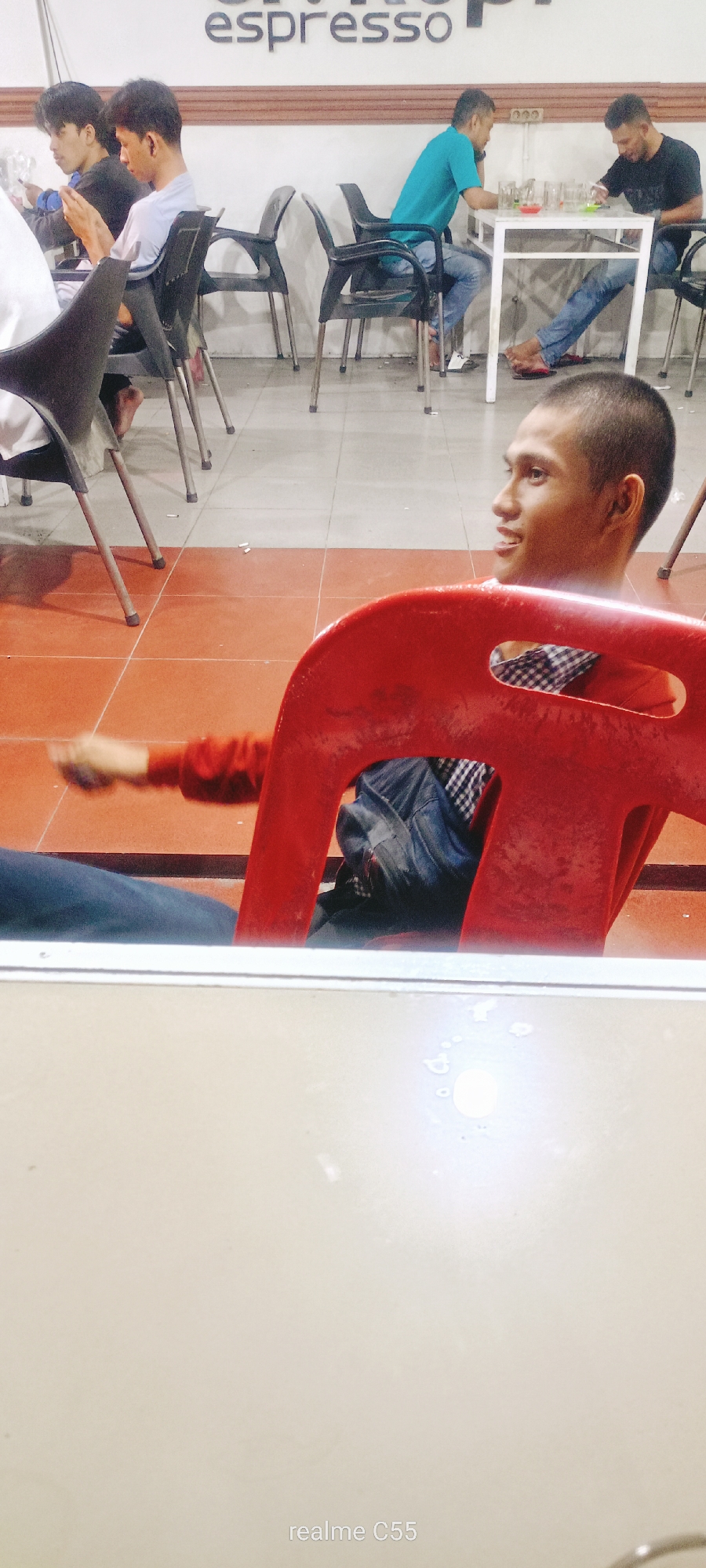 In Lhokseumawe, Indonesia, at a cafe, a young man with short hair sits backwards in a bright red plastic chair, looking to his right with a slight smile. He is wearing a plaid shirt and blue jeans. His left arm is extended forward, resting on the back of the chair. Behind him, partially visible, another person is also seated. In the background, several other individuals are seated at tables, with some using mobile phones and others interacting. One man in a blue t-shirt and jeans is seated at a white table, and another man in a black t-shirt and jeans is also at the table. The cafe has a tiled floor, with a checkered pattern of light and dark tiles. The word "espresso" is visible on a wall in the background. A white table is in the foreground, with some water droplets on its surface.