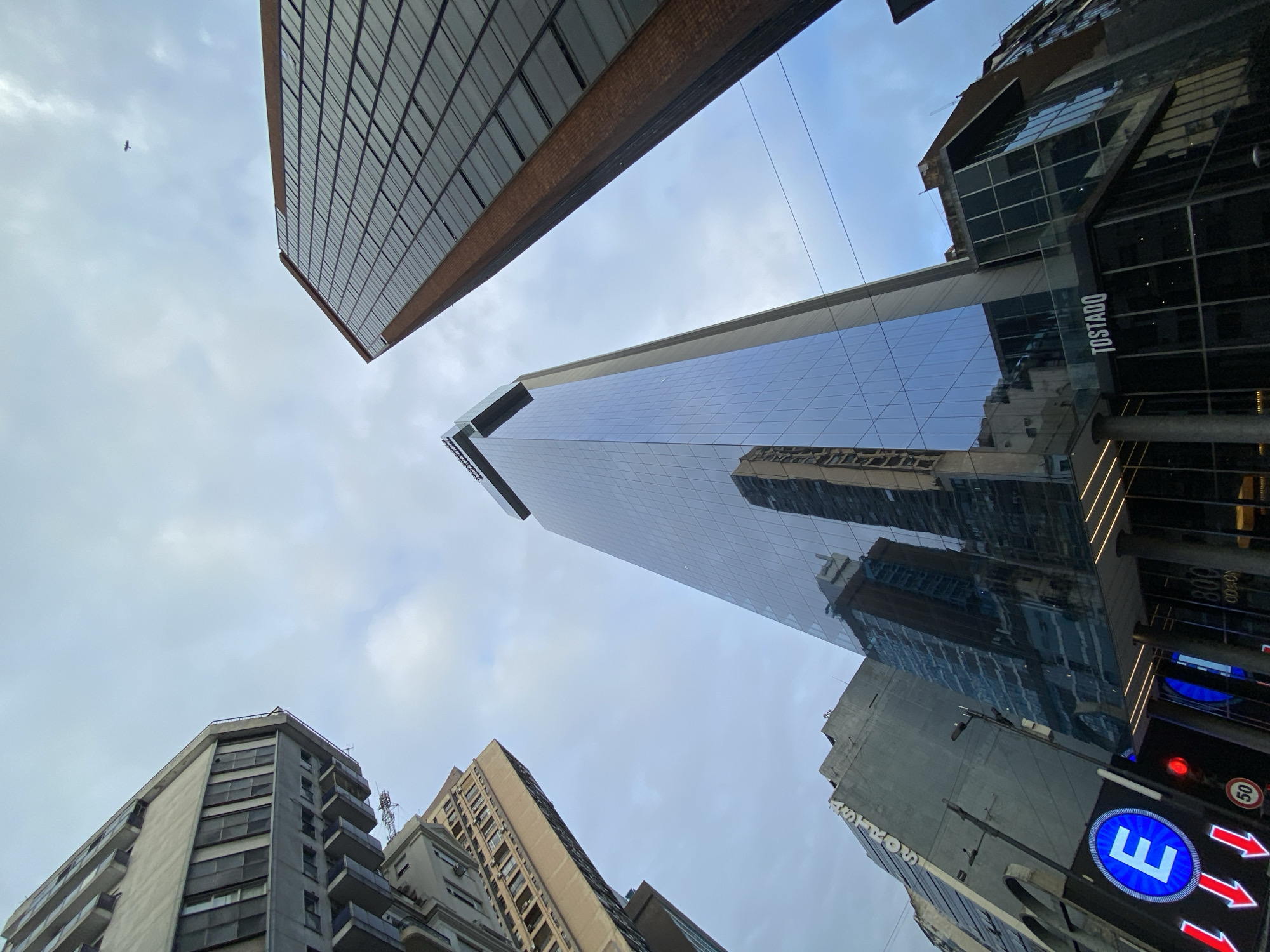 A low-angle, upward-looking photograph captures a cluster of tall buildings under a light gray, overcast sky. The central structure is a modern, slender skyscraper with a dark blue, reflective glass facade, identified by "800 ODON" and "TOSTADO" signage at its base. This building is located at **Corrientes 800, Buenos Aires, Argentina**, corresponding to Torre Odeon.
To the immediate left of the central skyscraper, an older, multi-story building with a light gray concrete facade, numerous rectangular windows, and small balconies is partially visible. Below this, another building section displays the partial word "ASTROS" arranged vertically on its upper facade. To the right of the central skyscraper, a tall building with a reddish-brown brick-like facade and a grid of dark windows extends upwards.
In the lower-left foreground, a circular blue street sign with a white capital "E" and three red downward-pointing arrows is visible. Adjacent to it, a red traffic signal and a black street lamp pole with multiple light fixtures are present. The sky is largely covered by uniform light gray clouds. A single, small, dark avian-like object is visible in flight in the upper-right portion of the sky. No individuals are discernibly present within the scene.