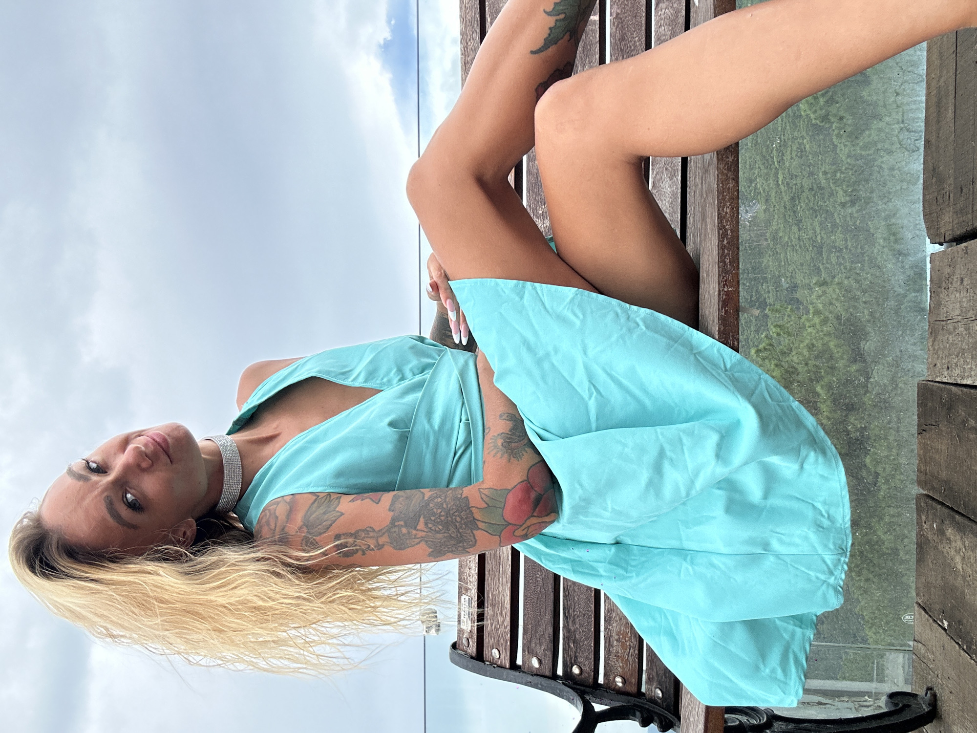 A blonde woman with tattoos is sitting on a wooden bench outdoors. She is wearing a teal dress and a sparkly choker necklace. The background shows a cloudy sky and a blurry green landscape.