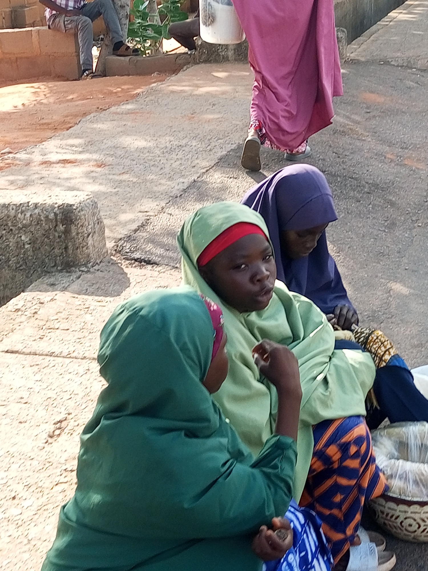 In Tempure, Nigeria, three individuals are visible, two of whom are partially obscured. The foreground features a young person wearing a green headscarf and garment, with a red band visible underneath. This individual is looking towards the camera with an open mouth. Behind them, another person is wearing a purple headscarf and appears to be engaged in an activity with their hands. To the right of these two, a portion of a third individual is visible, wearing a pink garment and a white bucket. They are walking away from the camera. The setting appears to be outdoors, with a textured ground composed of sand and gravel. In the background, a person is seated on a raised brick structure, wearing a plaid shirt and grey trousers. The overall scene suggests a public space, possibly a street or market area.