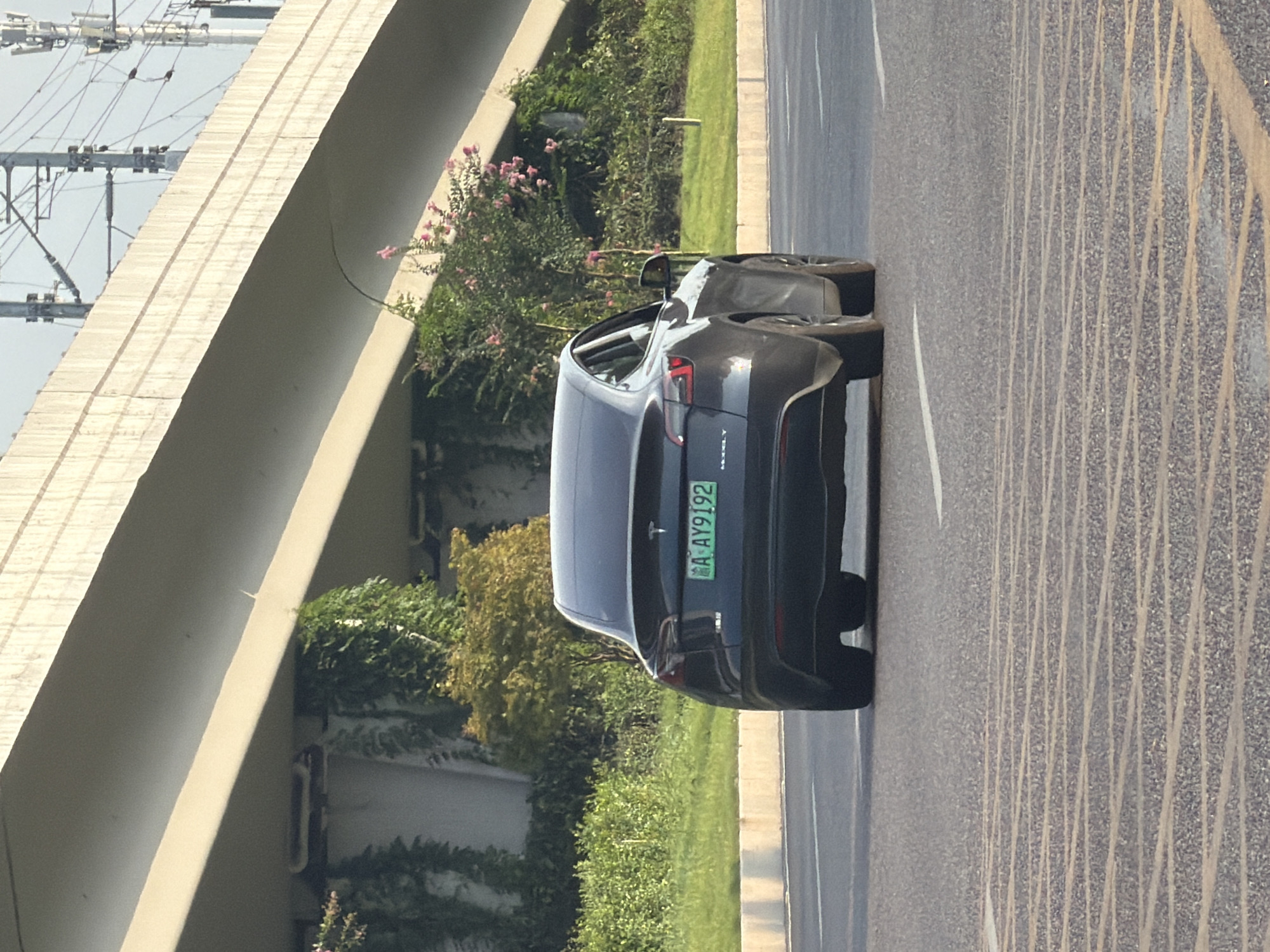 A dark gray Tesla Model Y is driving on a paved road in Jiangjin District, Chongqing, China. The car has a green license plate that reads "A AY9192". The vehicle is seen from the rear, with its taillights illuminated. The road has lane markings and the surrounding environment includes a concrete overpass with vines and bushes growing underneath and beside it. In the background, there are power lines and poles, suggesting an urban or suburban setting. The lighting indicates it is daytime, possibly late afternoon due to the shadows.