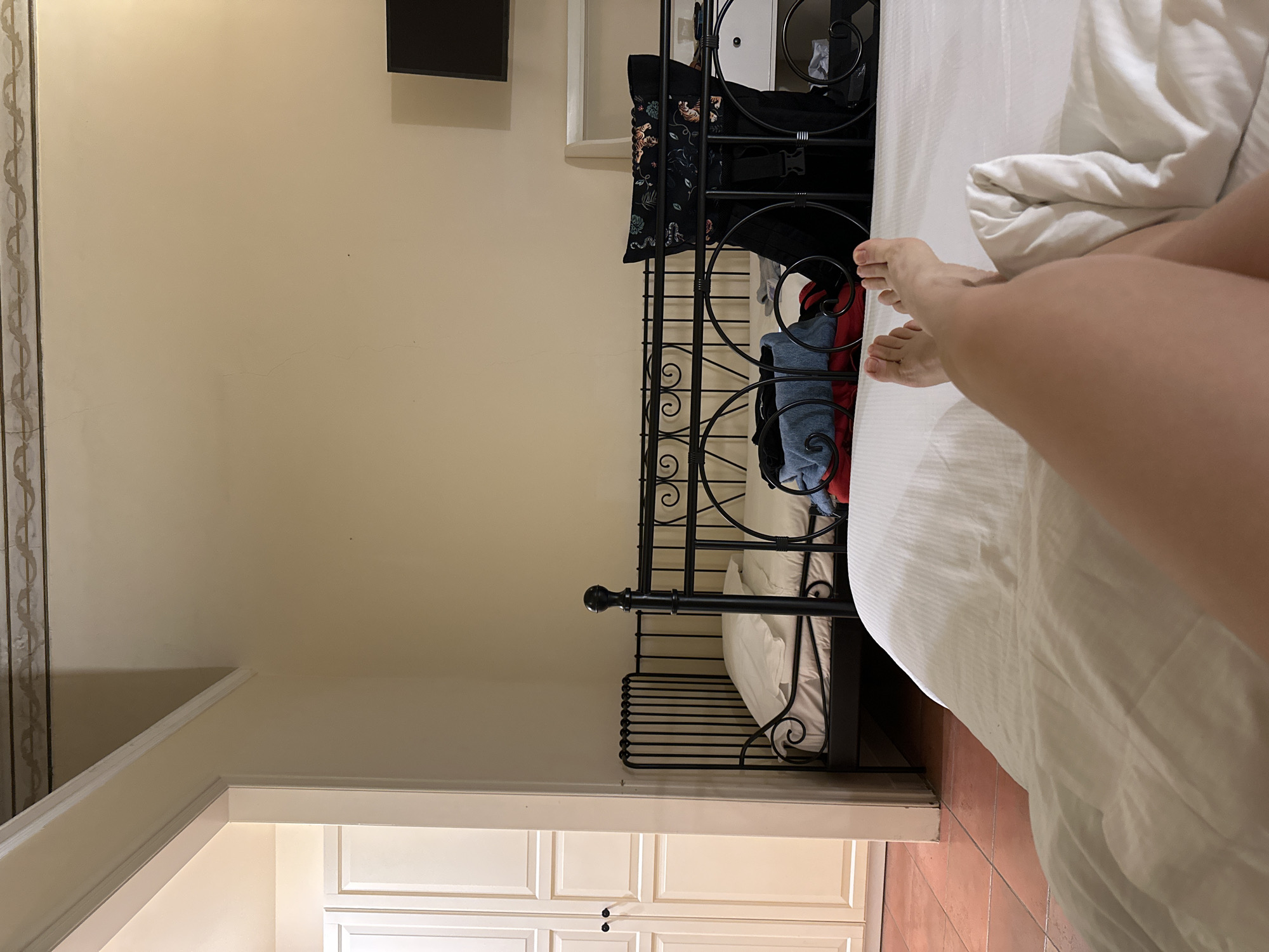 The image depicts a bedroom interior. The foreground shows a person lying in a bed with white linens. Their feet are visible, extending towards the center of the image. To the left, a black wrought-iron bed frame is visible, with some clothing and a bag hanging off the side. In the background, there's a light-colored wall with a mounted television. A partially visible doorway leads to a room with white cabinets. The floor is tiled with red terracotta tiles. The image suggests the location is likely Catania, Italy, given the architectural style and decor.