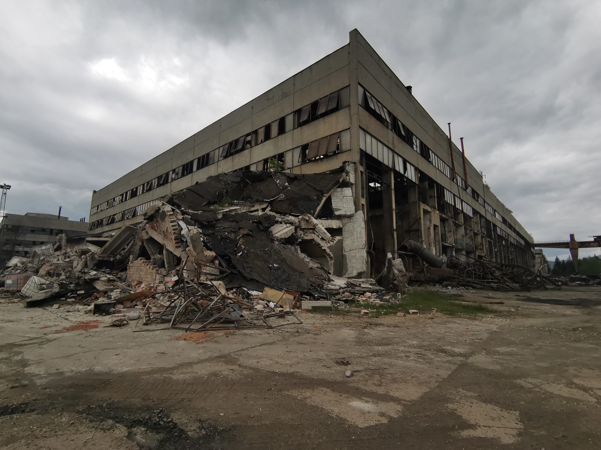 The image shows a partially collapsed, multi-story industrial building in Zhivkovo, Bulgaria. The building is made of concrete and has numerous broken windows. A large pile of rubble, including brick, metal rebar, and concrete debris, is in front of the building. The area in front of the building is paved with concrete and dirt. The sky is overcast. A crane is visible in the background to the left of the building, and a bridge or gantry is visible in the background to the right of the building.