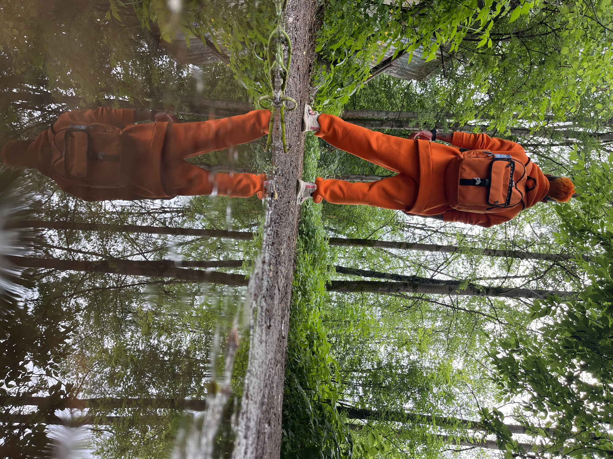 An eye-level, exterior shot depicts a person in an orange sweatsuit standing in front of a body of water. The person's reflection is visible in the water. The scene is surrounded by green trees. The location is identified as the city of Chaikovsky, Russia. A dirt path runs between the person and the water.