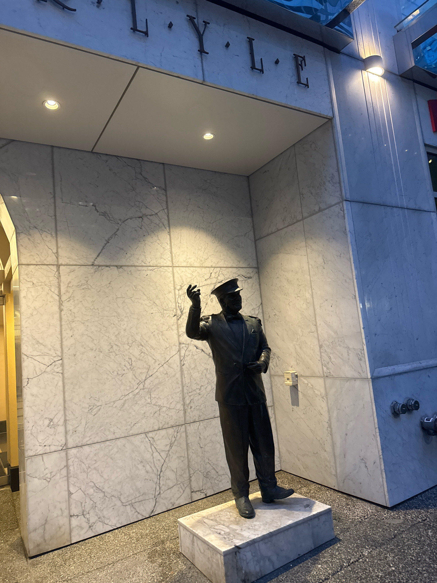 The image captures a bronze statue of a man in a uniform, possibly a military or police officer, standing on a marble pedestal outside a building. The statue is dressed in a hat, jacket, and trousers, with one arm raised as if gesturing or commanding. The background features a building with a marble facade and signage that reads "L.LYLE". The lighting suggests it is either dusk or dawn, with artificial lights illuminating the scene. The statue is positioned in an alcove-like entrance, with overhead lights and wall sconces providing illumination. Visible on the right side of the building are fire department connections. The ground surface is paved. The overall impression is one of public art displayed at the entrance of a building. The signage "L.LYLE" suggests a potential connection to a specific person or establishment. A search for "L. Lyle Vancouver" reveals this statue is located at the Fairmont Hotel Vancouver, and the statue depicts Colonel Lyle, a former manager of the hotel.