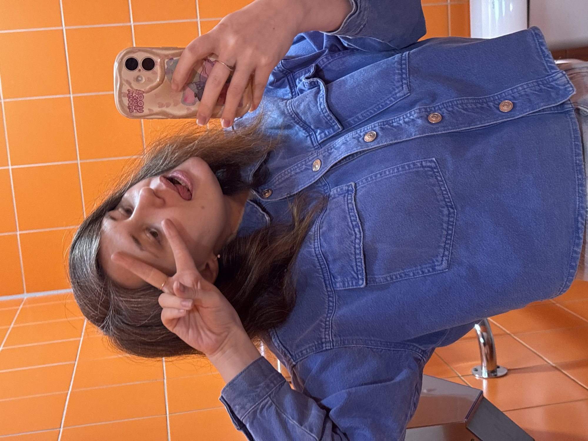An adult female is depicted taking a mirror selfie in a restroom located in Izhevsk, Russia. She has shoulder-length dark brown hair with visible graying at the roots. She is wearing a medium-wash blue denim long-sleeved shirt featuring gold-colored buttons and two chest pockets. Her left hand is raised, forming a "peace" sign, while her right hand holds a light-colored smartphone encased in a transparent, decorated cover displaying small heart graphics and inverted text. A gold-toned ring is present on her right ring finger. Her head is tilted, and her tongue is slightly extended. The background consists of bright orange square wall tiles with white grout lines. Visible restroom fixtures include a dark gray hand dryer and a chrome grab bar on the left, and a portion of a white porcelain fixture, likely a toilet or sink, on the right.