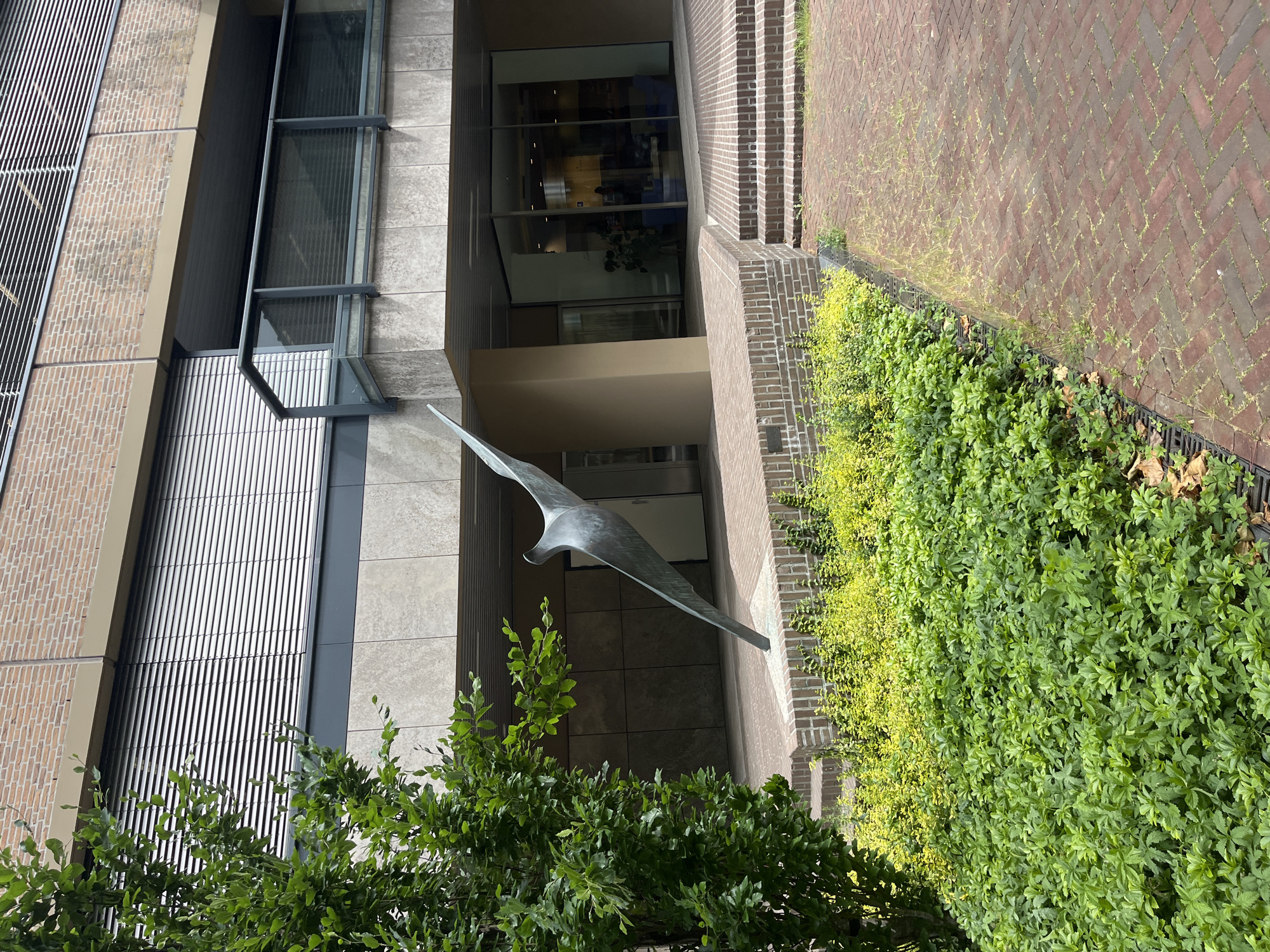 An outdoor, eye-level, medium shot captures a bronze abstract sculpture resembling a bird in flight. The sculpture is positioned in front of a modern building with a brick facade and large glass windows. Lush green bushes and yellow foliage surround the sculpture, and a red brick pathway leads towards the building's entrance. The lighting is natural, suggesting daytime. The scene appears to be a public space, possibly a plaza or courtyard.