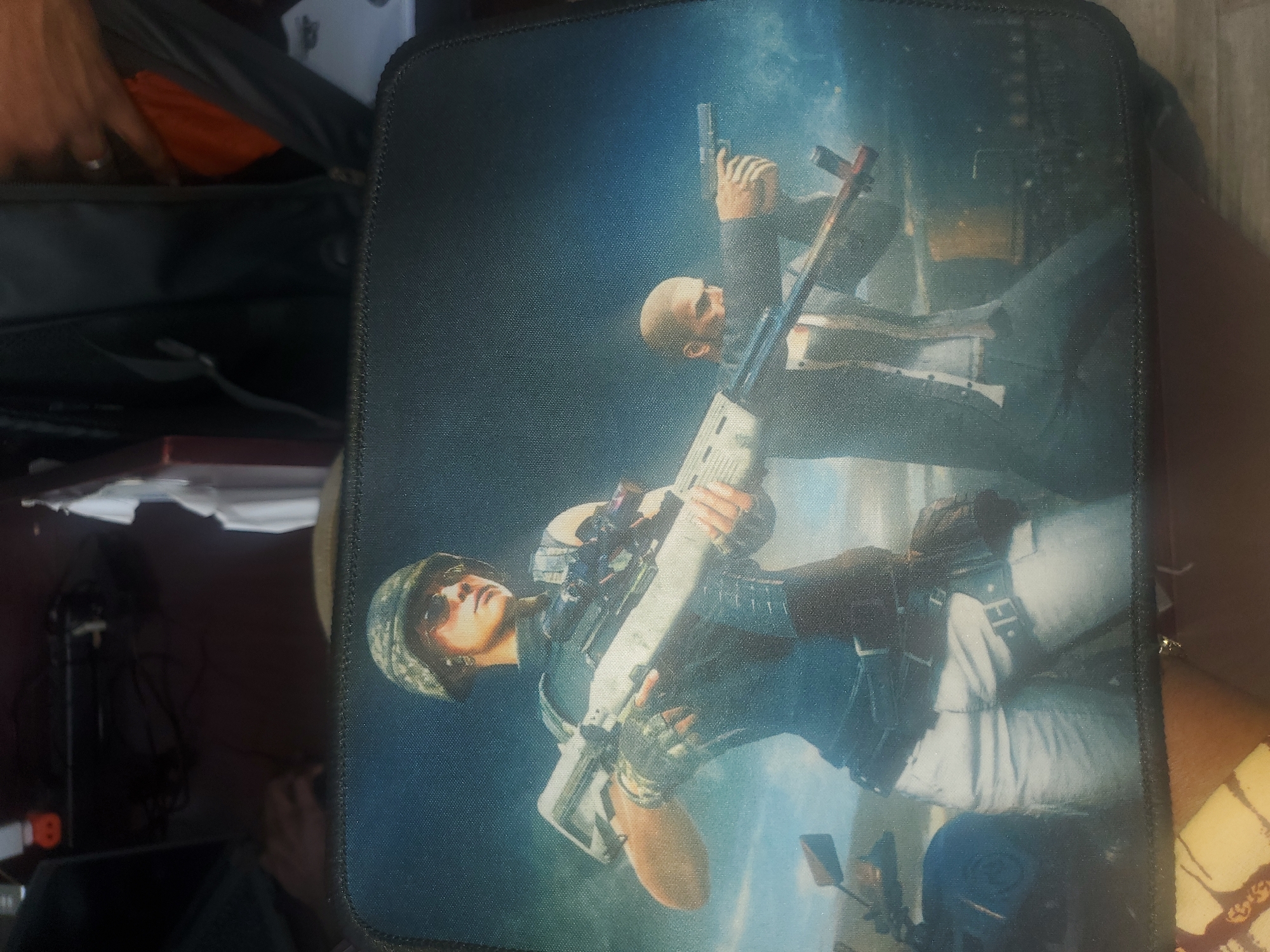The image is a close-up, overhead shot of a mousepad featuring a digital illustration of two figures in a combat-like setting. The foreground figure, on the left, is a woman wearing a camouflage helmet, sunglasses, and a tactical vest over a dark shirt. She is holding a large rifle with a scope. Her lower body is clad in white pants with tactical belts and holsters. To the right and slightly behind her is a bald man in a dark suit and white shirt, aiming a pistol. Both figures appear to be in action, with a background suggesting an urban or industrial environment with a hazy sky. There is also a motorcycle visible on the left side of the image. The mousepad itself has a black stitched border. The image is likely a depiction of characters from a video game. The scene does not depict any recognizable landmarks or features of Gwagwada, Nigeria.