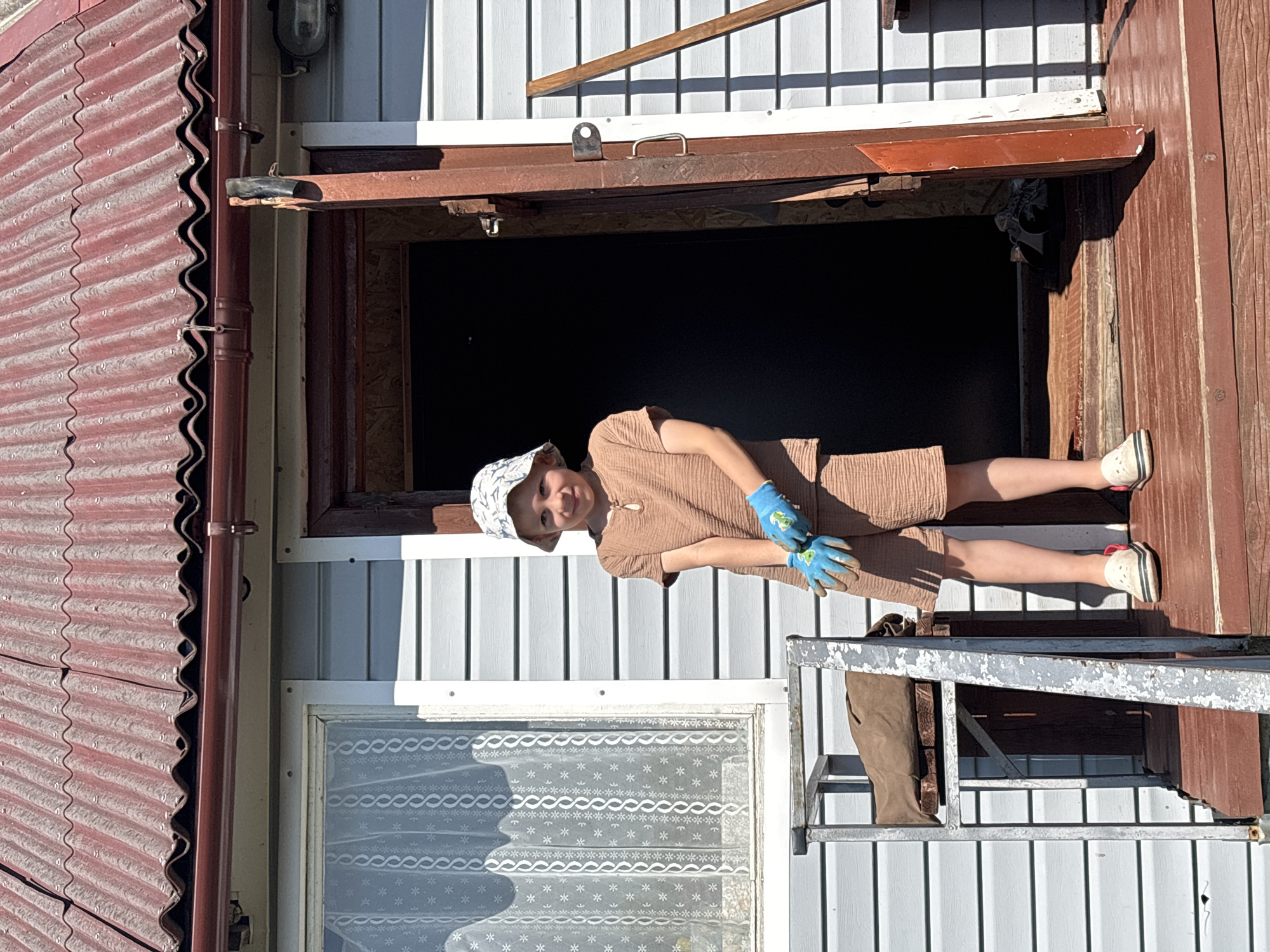 A young boy wearing a tan t-shirt, matching shorts, a bucket hat, blue gardening gloves, and white sneakers stands on a wooden porch in front of a light blue clapboard house with a red corrugated roof. He is holding gardening tools in his gloved hands. The door to the house is open, revealing a dark interior. To the left of the boy is a window with a white lace curtain. A metal railing with a brown canvas bag is visible on the porch. The scene appears to be in a rural setting, possibly in the village of Dunilovo, Russia, given the architectural style and weathered appearance of the structures.
