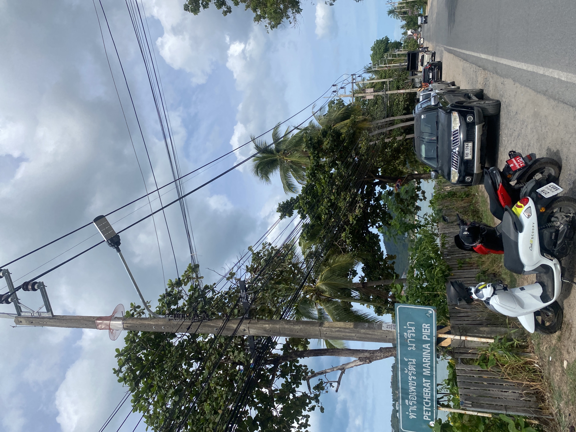 The image captures a coastal road scene in Baan Bang Rak, Thailand, under a partly cloudy sky with visible blue patches. In the immediate foreground on the left, two scooters are parked on a dirt shoulder adjacent to a wooden fence. The white scooter on the left is a Honda Club Scoopy, displaying a partial Thai license plate "ช 4487", with a black helmet resting on its seat. To its right, a red and black scooter shows two Thai license plates: a red plate "บษ 7101" and a standard plate "2กณ 5857".
Behind the scooters, a rectangular green sign with white text reads "ท่าเรือเพชรรัตน์ มารีน่า" and "PETCHERAT MARINA PIER". Beyond the wooden fence and sign, a body of water, likely a bay or ocean, is partially visible through dense tropical foliage. Distant hills are discernible across the water.
A tall, weathered concrete utility pole stands prominently in the mid-ground, heavily equipped with numerous black electrical and communication cables. It features multiple insulators and a solar-powered street light extending horizontally to the left. A reddish-pink plastic sheath encircles the pole below the uppermost insulators. Surrounding the utility pole and extending across the left side of the image are abundant tropical trees, including several tall palm trees (likely coconut palms) and large leafy trees, which partially obscure the view of the coastline.
On the right side of the image, a paved asphalt road extends into the distance, curving slightly right. A black SUV, identified by its Thai license plate "กข 1764", is parked on the dirt shoulder to the right of the utility pole, facing the viewer. Further down the road, a line of parked vehicles includes a silver pickup truck, a dark sedan, and a white truck. Additional utility poles and dense vegetation line the roadside as it recedes into the background. No individuals are present in the scene.