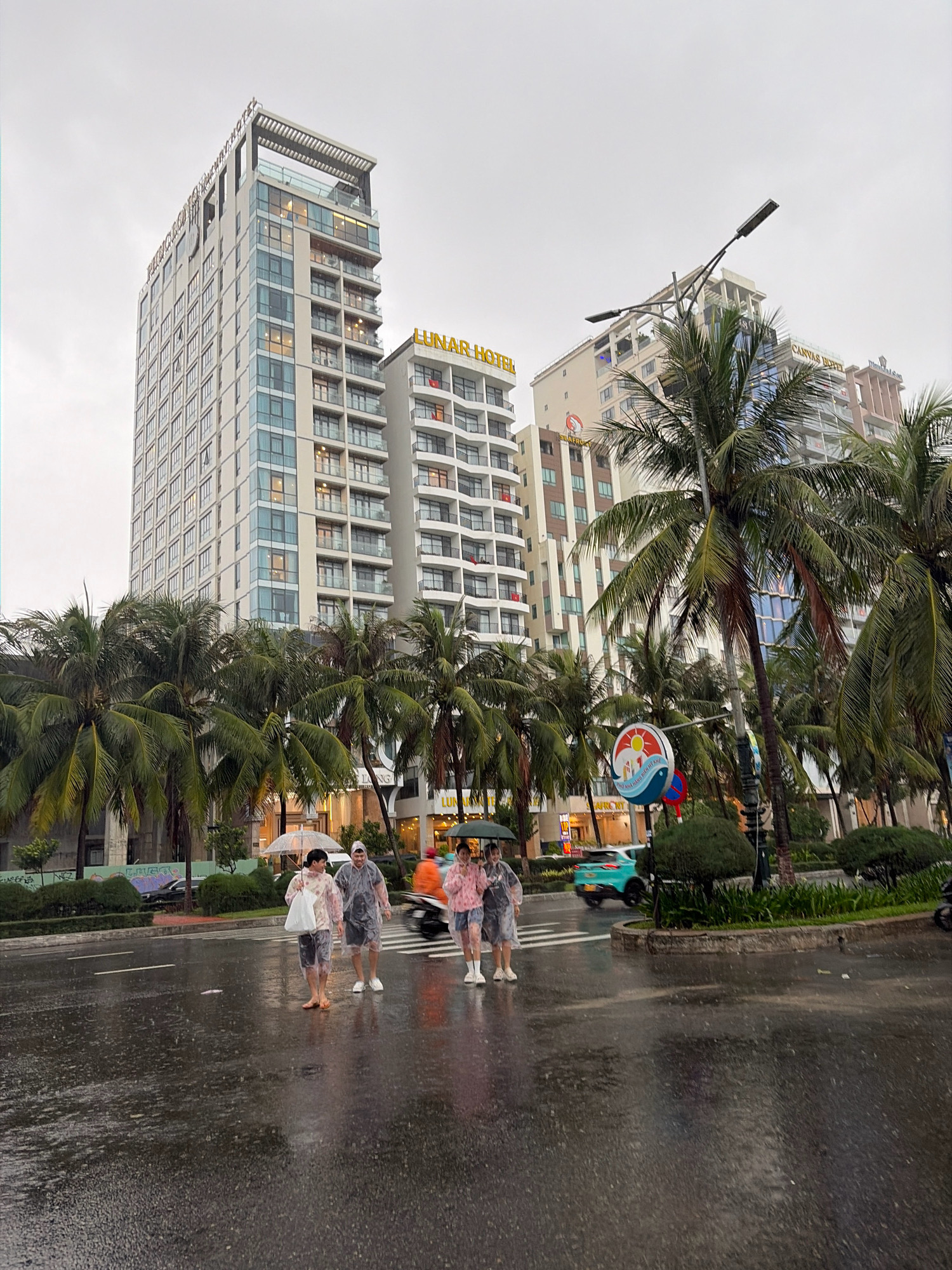 A group of three people are walking across a wet street in Da Nang, Vietnam, during a rain shower. They are wearing clear and pink raincoats and one person is holding a clear umbrella. The street is slick with rain, and palm trees line the sidewalk. In the background, several tall buildings, including the Lunar Hotel, are visible. A turquoise car is driving by on the street, and a motorbike is parked on the right side of the image. The sky is overcast and gray.