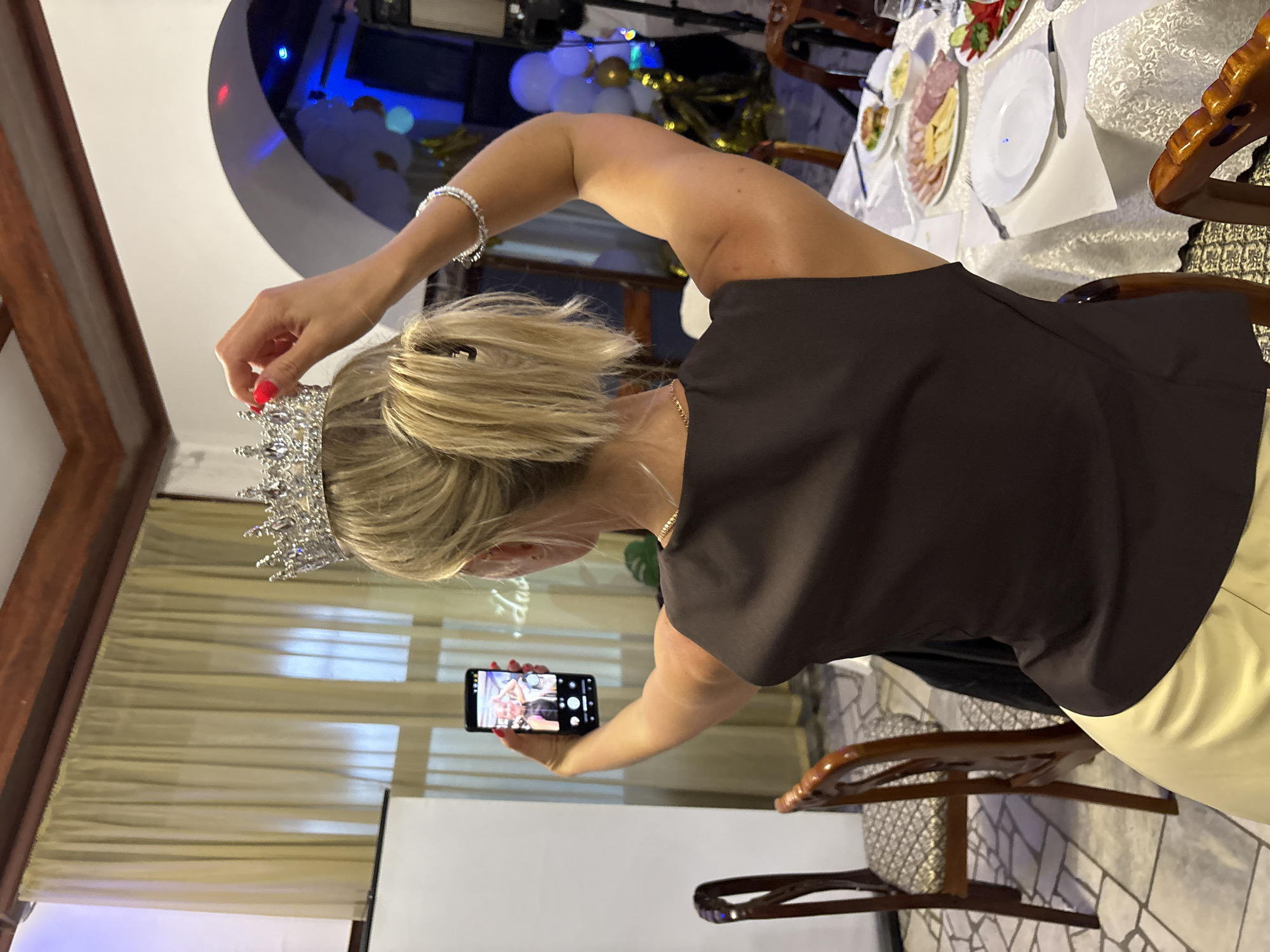 A woman with blonde hair is holding a tiara to her head while taking a selfie with her phone. She is wearing a dark top and a light-colored skirt. In the background, there are balloons and a table with food, suggesting a celebration or party. The lighting is dim, but there is some blue and red lighting from what appears to be decorative lights. The scene looks like an indoor event.