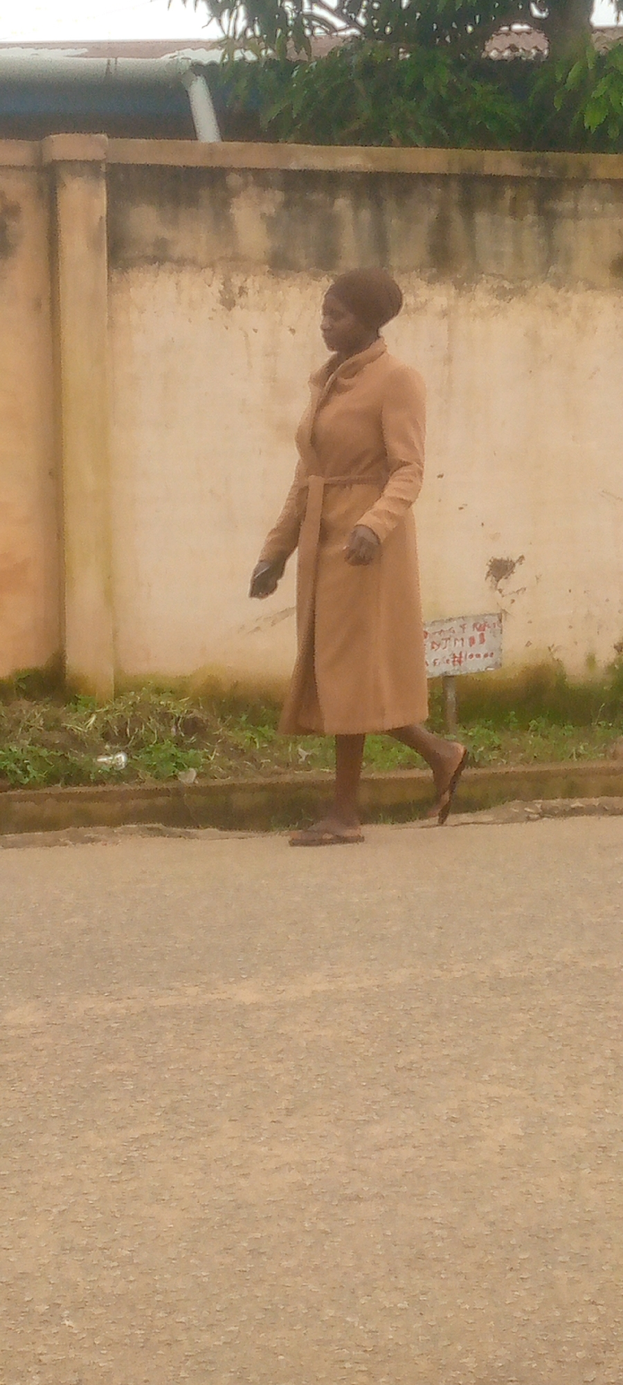 A full-shot photograph captures a young woman walking in profile from left to right on an asphalt road. She is wearing a tan trench coat with a belt cinched at her waist and matching headwrap. Her feet are bare except for simple, flat sandals. The woman is walking on the road, with a patch of green grass and weeds bordering it. Behind the grass is a light brown, textured wall. Trees with green foliage are visible above the wall, suggesting an outdoor environment with natural elements. The overall lighting suggests it is daytime.