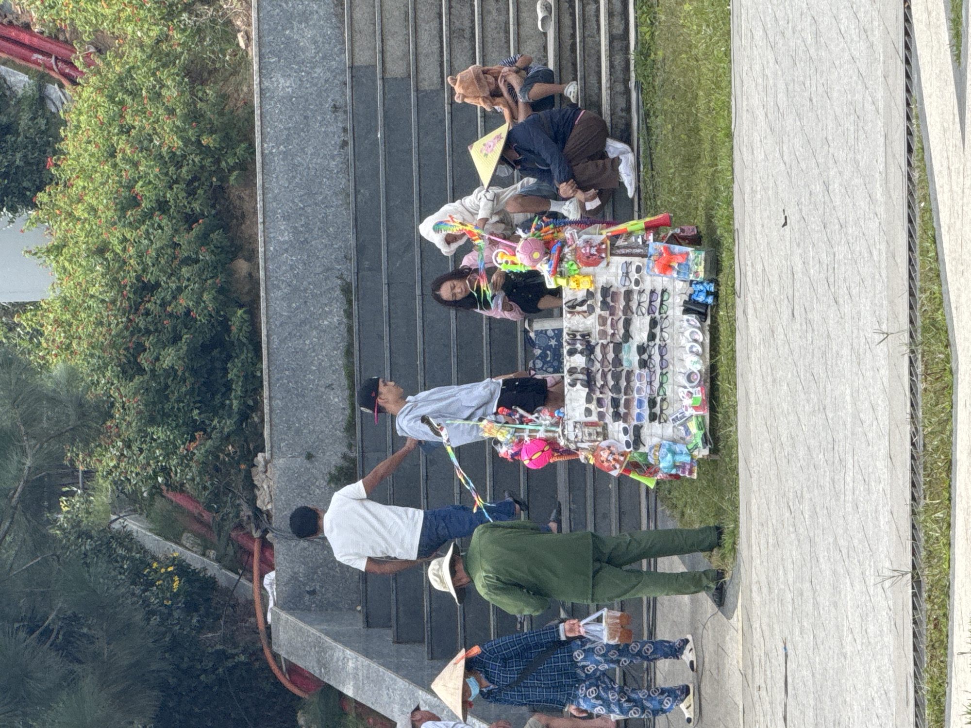 In Da Lat, Vietnam, on a bright, sunny day, a street vendor stands by a cart filled with sunglasses and toys on a paved walkway at the base of a wide, grey stone staircase. Several people are seated on the stairs. A person in a green uniform, likely a vendor, stands near the cart. A woman wearing a conical hat and a blue plaid shirt is walking past. Further up the stairs, two young men, one in a white t-shirt and the other in a grey hoodie, are standing. Another group is seated on the stairs, including a woman in a pink shirt, a person in a white hooded outfit, and a child in a brown fuzzy hat. The environment features lush green bushes and trees, suggesting an outdoor public space, possibly a park or a tourist attraction.