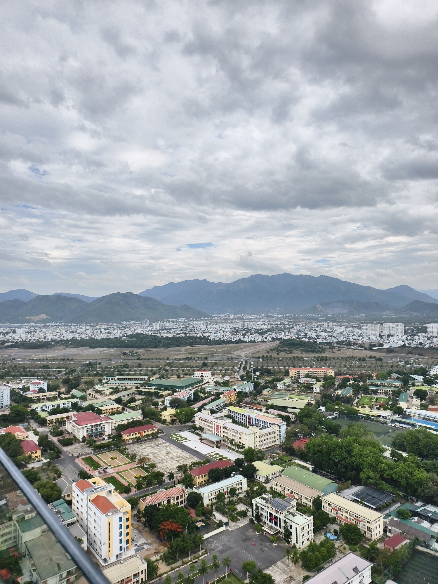 This aerial photograph captures a sprawling cityscape nestled against a backdrop of distant mountains under an overcast sky. The view is from a high vantage point, looking down upon a mix of urban and natural elements.
In the immediate foreground, a dense collection of buildings is visible, characterized by varied architectural styles. Many structures are low-rise, with some featuring traditional red or orange tiled roofs and others having flat, grey or green roofs, indicative of institutional or industrial facilities. There are extensive paved areas, green courtyards, and tree-lined streets, suggesting a well-planned complex, possibly a school, university, or government compound. A prominent multi-story building with white walls and blue window accents stands out, partially obscured by what appears to be a railing or part of the viewing platform.
The midground reveals further urban expansion, with numerous white and light-colored buildings forming a dense residential or commercial area. These structures extend towards a wide, undeveloped stretch of land that appears to be a dry riverbed or a large, cleared area, separating the foreground development from the more distant city. This section is dotted with sparse vegetation and what look like roads or pathways.
In the far background, a majestic range of mountains rises, their peaks appearing hazy and dark blue-green under the heavy cloud cover. The sky is entirely overcast, filled with thick, grey clouds that diffuse the sunlight, creating a soft, even illumination across the landscape. The overall weather appears calm but subdued.
No specific human figures are discernible, and no particular event is taking place, presenting a static snapshot of the urban-natural interface. There is no visible text in the image. The scene suggests a daytime setting in an area experiencing significant urban development, likely in a region with prominent natural features like mountains.