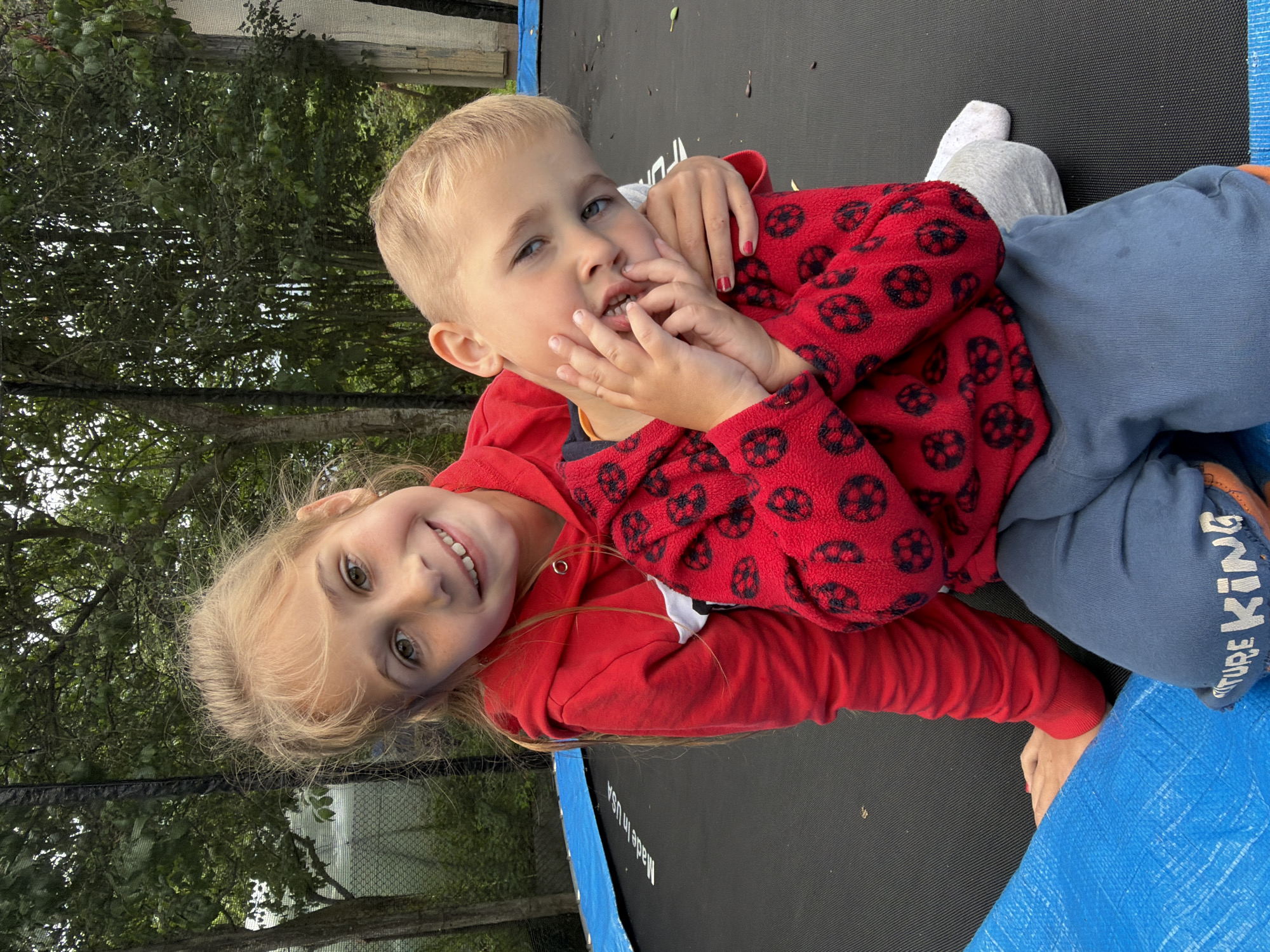 Two children, a girl and a boy, are sitting on a trampoline in an outdoor setting, possibly a backyard. The girl, with blonde hair and a red hoodie, is embracing the boy, who is wearing a red top with a ladybug pattern and blue pants. The boy is making a playful gesture with his hands near his face. The trampoline has a blue safety mat along the edge and a black jumping surface. In the background, there are trees and foliage, suggesting a natural environment. The overall scene captures a moment of playful interaction between the two children.