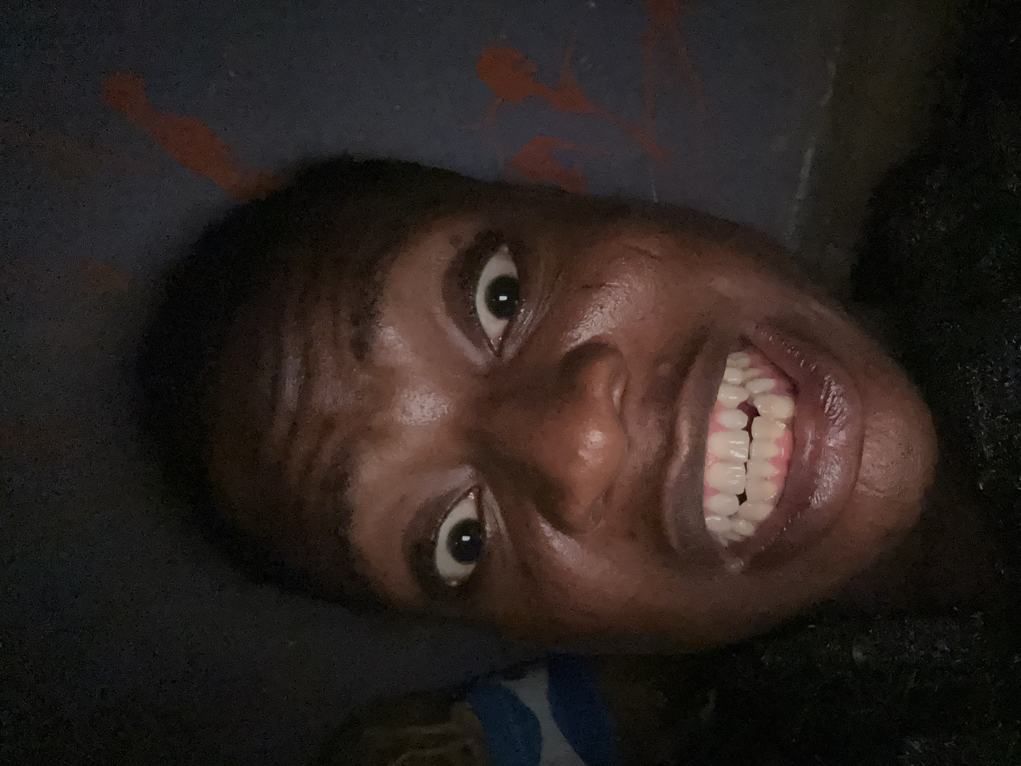 A close-up, slightly angled shot of a person's face. The person has dark skin, dark hair, and is smiling, revealing their teeth. The background is dark, with some indistinct orange markings visible above the person's head. The setting and location are Unknown, Unknown.