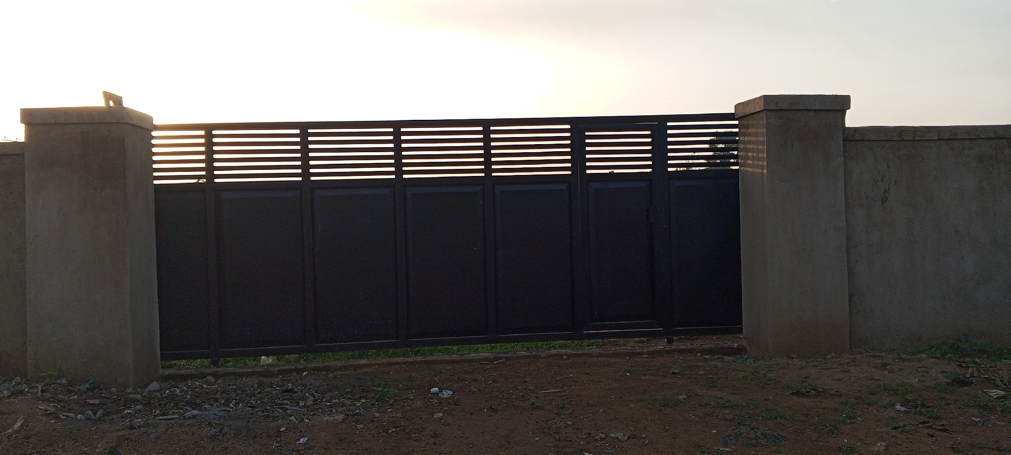 This is a wide, eye-level, outdoor, natural light photograph of a dark-colored, metal gate with horizontal slats across the top, set between two concrete pillars. The gate is positioned in front of a concrete wall. The ground in front of the gate is covered with dirt, small stones, and scattered debris. Green grass is visible beneath the gate. The sky is bright and overcast with the sun setting or rising, creating a bright haze. The image appears to be taken in Jalingo, Nigeria, given the context.