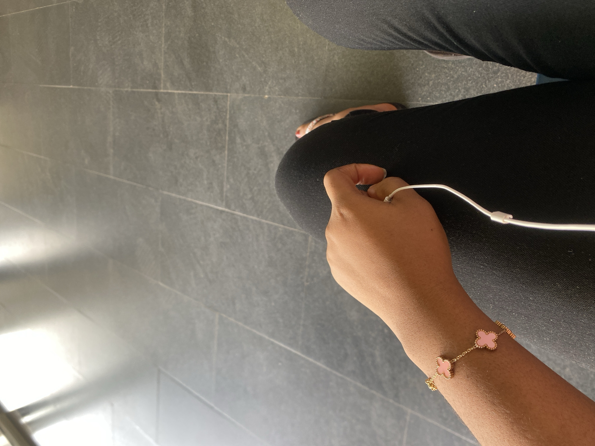 The image depicts a person sitting with their legs crossed, wearing black leggings and a gold bracelet with pink clover-shaped charms on their right wrist. They are holding a white charging cable in their hand. The floor is tiled with gray rectangular tiles. The location is unknown and cannot be determined to be in Abuja, Nigeria based solely on this image.