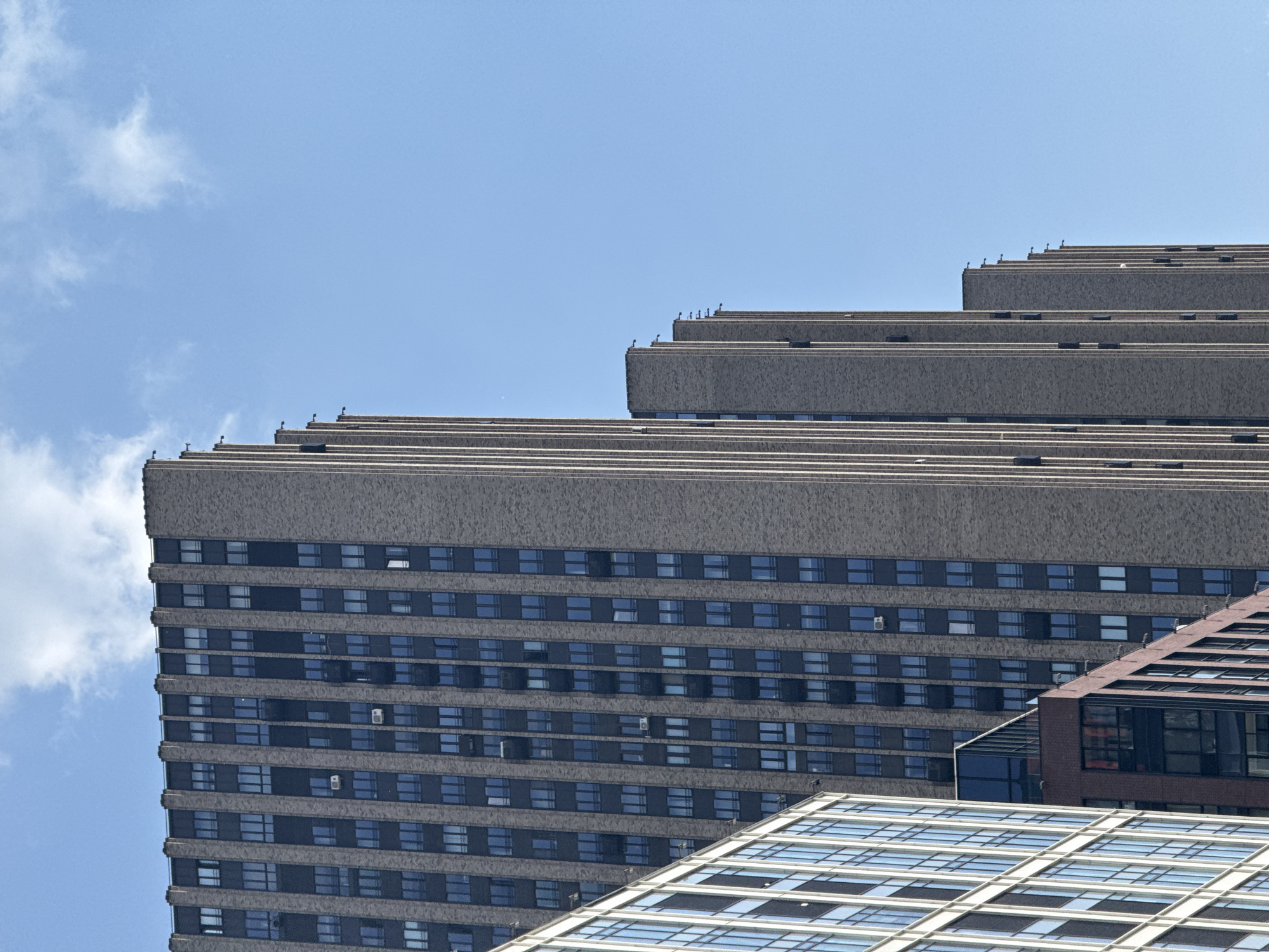 The image shows a clear blue sky with scattered white clouds above a series of modern high-rise buildings. The main building on the left is a tall, rectangular structure with a brick facade and numerous dark windows arranged in a grid pattern. Vertical dark bands, likely part of the window frames or structural elements, run down the face of the building, creating a ribbed appearance. To the right of this building, several other similar structures are visible, receding in size and perspective, also with brick exteriors and repeating window patterns. One building on the far left is different, appearing to have a lighter-colored facade with more glass. The overall impression is an urban landscape dominated by contemporary architecture.