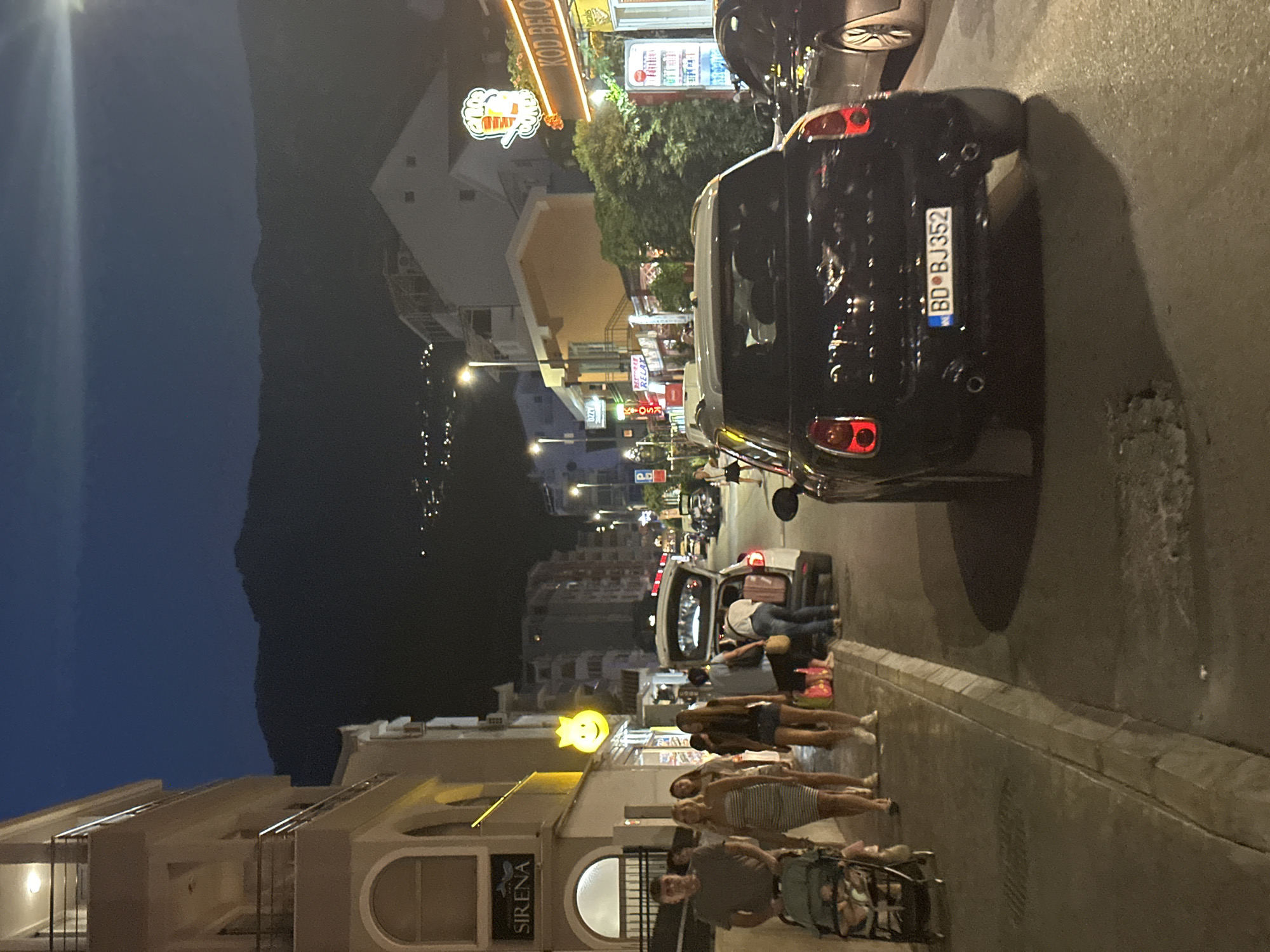 The image depicts a street scene in Budva, Montenegro, at dusk or early evening. A dark-colored Mini Cooper with license plate BD BJ352 is parked in the foreground, facing away from the camera. To the left, a sidewalk is occupied by several pedestrians. A man is pushing a stroller with a child. Behind him, a group of women are walking, one in a striped dress and another in a patterned dress. To the right of the sidewalk, a white SUV has its trunk open, and several people are loading or unloading items.
In the background, buildings line the street, some with illuminated signs. One sign reads "SIRENA". Further down the street, there are more buildings and streetlights, and a distant mountain range is visible against the dark sky, with scattered lights suggesting a settlement on its slopes. The overall atmosphere is one of a casual evening outing in a coastal town.