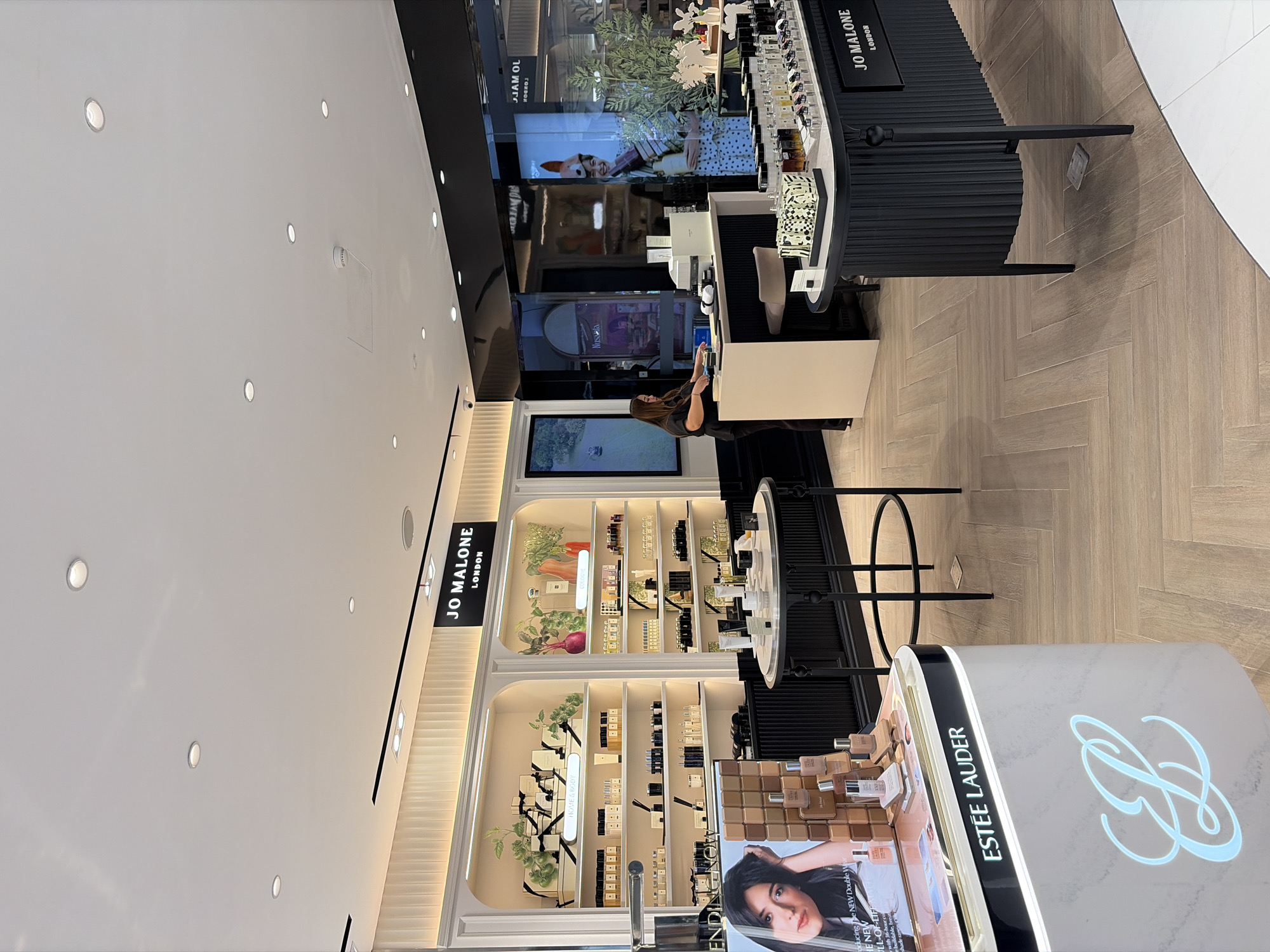 A well-lit interior scene depicts a high-end retail store, likely a department store beauty counter, located in Kuta, Indonesia. The store features a clean, modern aesthetic with white and black color schemes, accented by warm wooden flooring laid in a herringbone pattern. In the background, prominently displayed shelves are stocked with various beauty products and fragrances, including a section branded "JO MALONE LONDON" with accompanying signage. To the left, a display for "ESTÉE LAUDER" is visible, featuring a large advertising image of a woman and product samples. A woman in dark clothing is standing behind a counter on the right, appearing to be an employee interacting with the point of sale system. Several display tables with products are strategically placed throughout the store. The lighting is bright, suggesting an indoor retail environment with ceiling spotlights and illuminated display shelves.