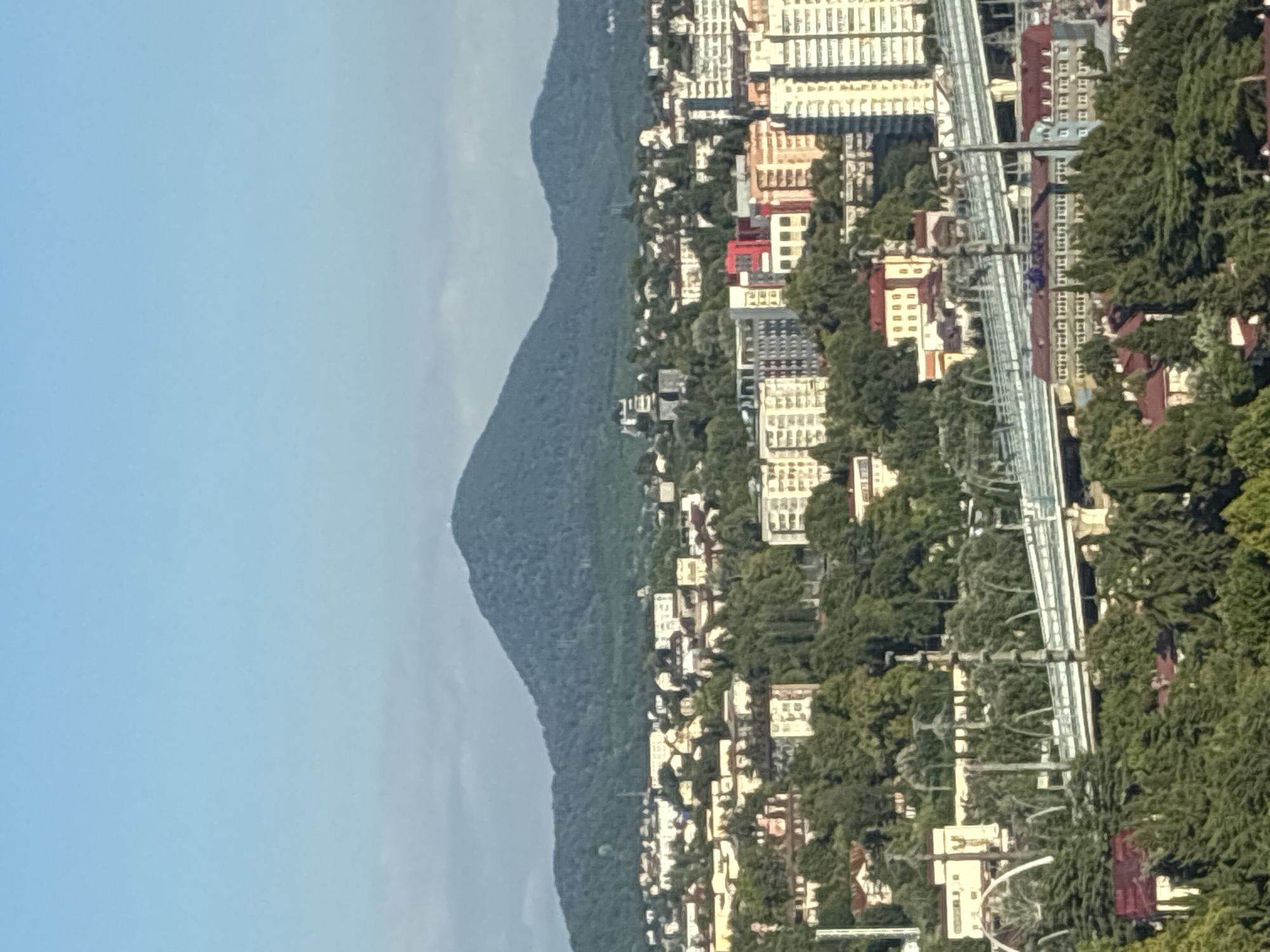 This is a daytime, high-angle shot of a city nestled in front of a large, tree-covered mountain. The sky is clear and blue, with some wispy clouds visible near the horizon. The city itself is a mix of low-rise buildings and taller apartment complexes, all surrounded by lush green trees. A multi-level bridge, likely an overpass or highway, dominates the mid-ground, with traffic visible on its surface. In the distance, the imposing mountain rises, its slopes densely forested. The scene suggests a vibrant urban environment integrated with a natural landscape. This is likely the city of Sochi, Russia, a popular resort town known for its coastal location and proximity to the Caucasus Mountains.