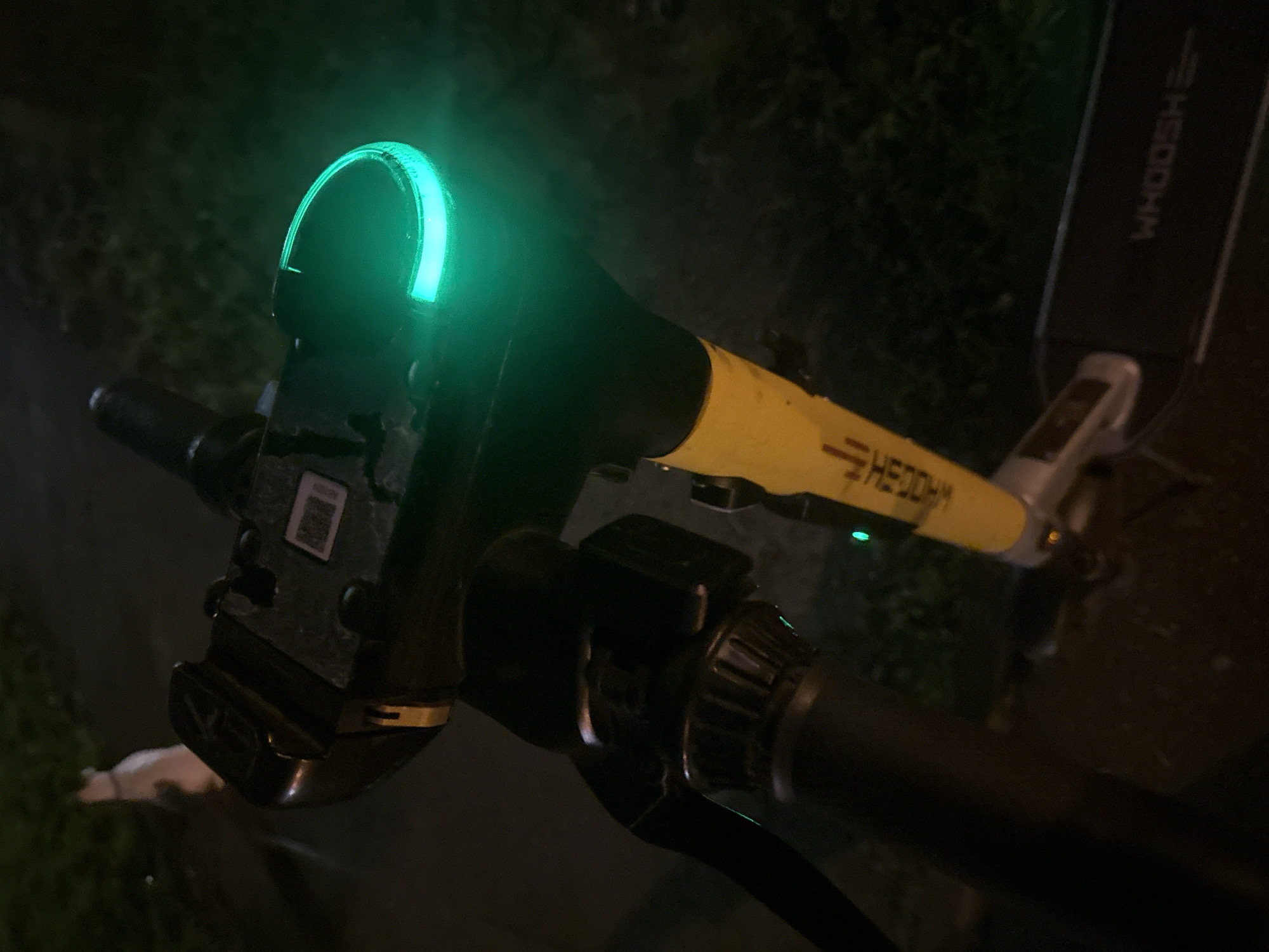 The image shows a close-up view of the handlebars and front section of a black and yellow electric scooter at night. A bright green light is illuminated on the front of the scooter, possibly indicating its status or a safety feature. The scooter has a QR code sticker on the upper black housing, and the word "WHOOSH" is visible in white lettering on the black deck of the scooter. The background is dark and out of focus, with some green foliage suggesting an outdoor setting, likely a sidewalk or park. No people are visible in the frame. The overall impression is that of a parked or momentarily stopped electric scooter in an urban environment.