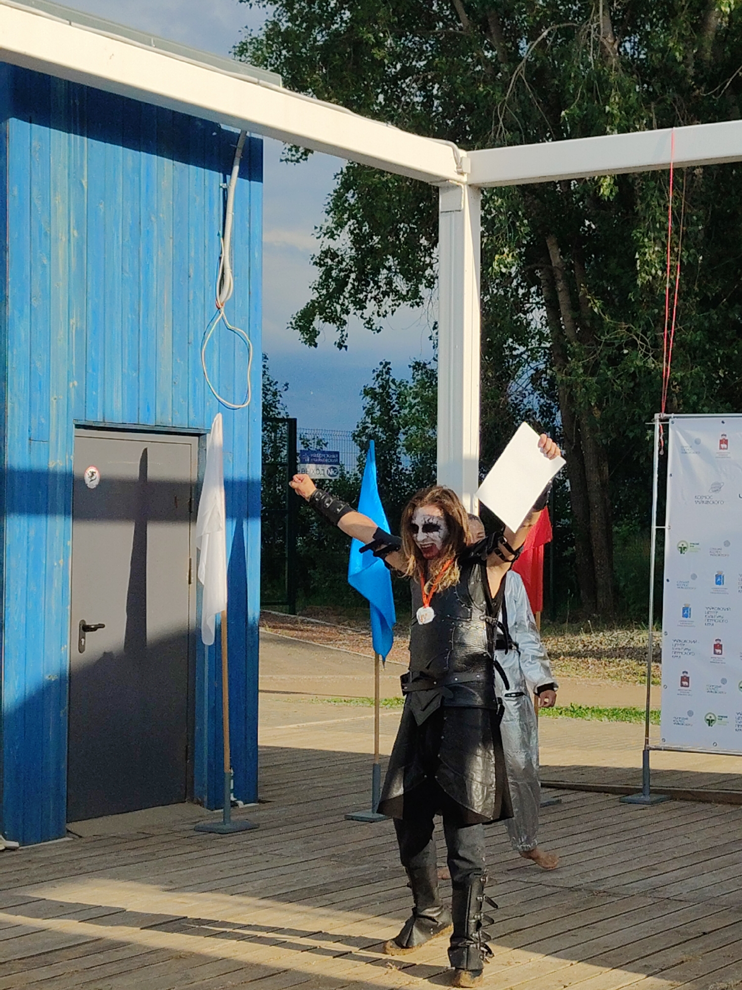In an outdoor setting, a person with elaborate white and black face paint, dark clothing resembling armor, and a medallion around their neck stands on a wooden deck, holding a white sheet of paper. Their right arm is raised, and they appear to be speaking or performing. Behind them, another person in a silver suit is visible. To the left is a blue wooden structure with a gray door, and a white flag on a pole. To the right, a white banner with text and logos is displayed. Trees and a fence are in the background under a cloudy sky.